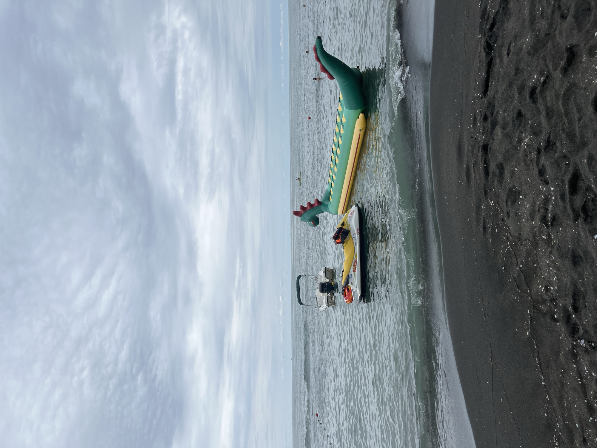 A view of the Black Sea coast in Ureki, Georgia, on a cloudy day. In the foreground, dark sand covers the beach with scattered white shells. Waves gently lap at the shore.
In the water, several objects are visible. A yellow jet ski with an orange life vest on it is positioned in the middle ground. To its right, a large, inflatable, green dragon-shaped water toy floats, its head and tail visible. Further back, a white boat with a green canopy is moored.
In the distance, the calm sea stretches to the horizon under a sky filled with overcast clouds. Several people can be seen in the water, some swimming and others in what appear to be kayaks. Red buoys are dotted in the water, marking a boundary. The overall scene suggests a recreational area by the sea.