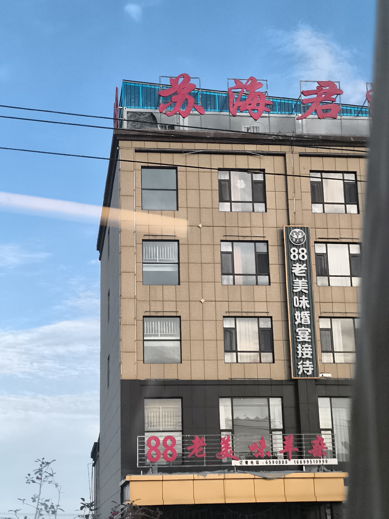 This is a daytime, eye-level, medium shot captured from outside of a building on a cloudy day. The building is tall and has multiple stories with several windows. The facade of the building is a light brown color with darker brown and black accents near the bottom. There are red Chinese characters on the top of the building and smaller red Chinese characters and numbers on the lower part of the building. A vertical sign with Chinese characters and numbers is attached to the building between some of the windows. There are electrical wires in the foreground. The sky is blue with scattered white clouds. This image appears to be taken in Lucaogou Town, China.