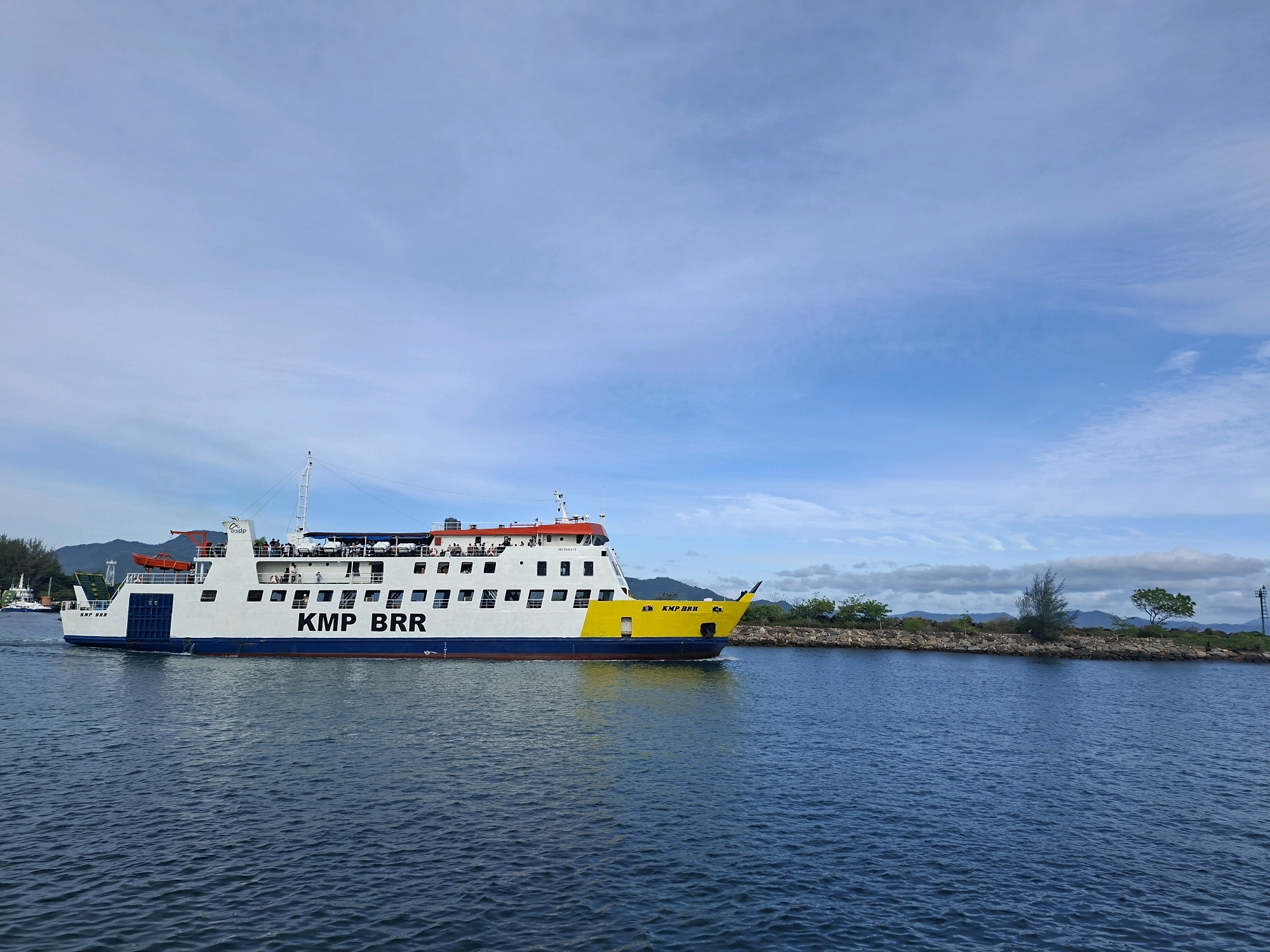 A large white ferry, identified by the lettering "KMP BRR" on its side, sails across a calm blue body of water. The ferry has multiple decks with numerous windows and a yellow bow. People are visible on the upper deck of the ferry. To the right of the ferry, a rocky shoreline is lined with sparse vegetation, including trees and bushes. In the background, a range of mountains is visible beneath a cloudy blue sky. The scene appears to be a coastal area. This location is identified as Banda Aceh, Indonesia, a city known for its coastal geography and maritime activity.