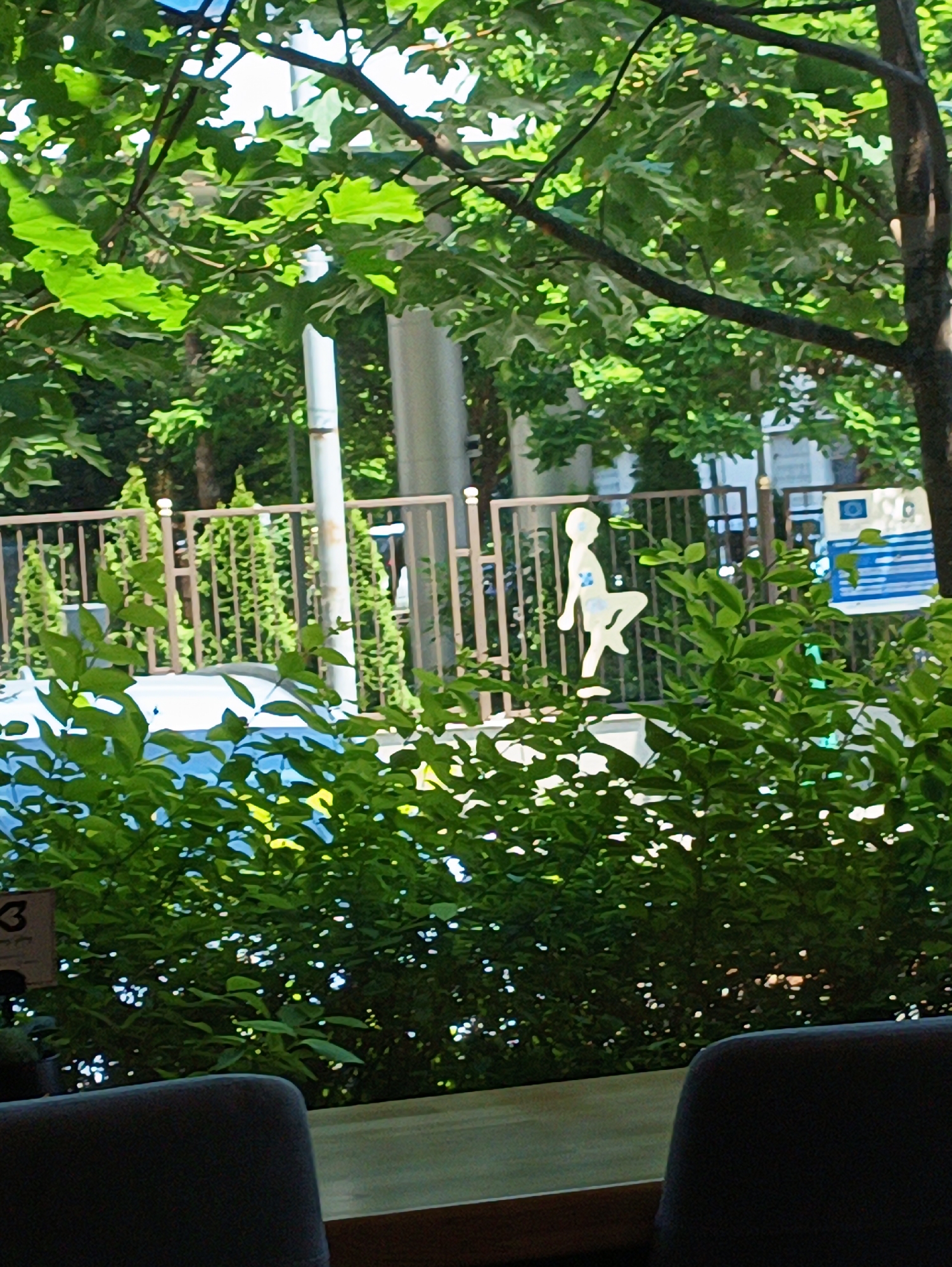 The scene, located in Burgas, Bulgaria, is an outdoor environment viewed from a shaded perspective, likely a café or restaurant. In the immediate foreground, the top of a dark chair back is visible in the bottom right, and a light-colored tabletop or counter extends from the bottom center. A partially obscured dark object with the number "3" is present in the bottom-left corner. A dense layer of vibrant green foliage occupies the lower and mid-ground. Behind these bushes, a stylized, white, anthropomorphic figure, resembling a child standing on one leg with the other bent upwards, is prominently positioned. A blue marking is visible on the figure's upper leg area. Behind the figure, a light-brown metal fence with vertical bars extends across the mid-ground. Beyond the fence, additional green foliage, a tall light-colored pole, and a partially visible blue and white sign with an accompanying blue bench are discernible. The background features a dense canopy of green trees with visible branches, through which patches of bright blue sky are apparent. The lighting indicates direct sunlight, creating bright highlights and deep shadows on the foliage. No living individuals or explicit human activity are present in the image.