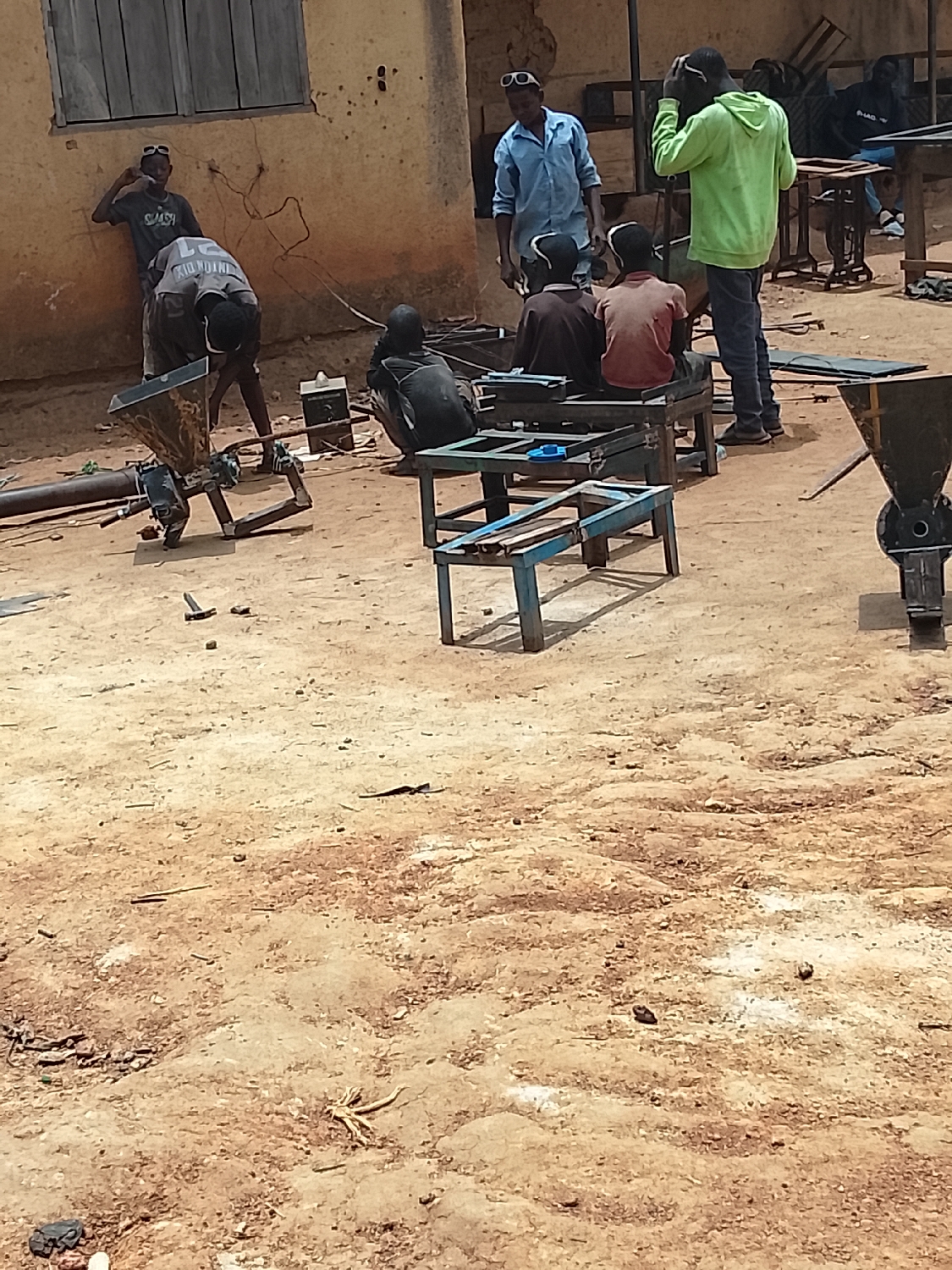 The image depicts an outdoor metalworking or fabrication workshop under bright daylight, set against a backdrop of simple, light-colored buildings, one featuring a wooden window shutter. The ground is uneven dirt with scattered debris.
At least seven males are present, engaged in various activities. On the left, one individual, wearing a shirt with "KIDUNDUNI" and possibly "21" visible, is bent over, seemingly working on a piece of machinery that includes a funnel-like component. Nearby, another person in a shirt with "SWASH" leans against the building. In the center, a person in a black shirt crouches near what appears to be a power tool or welding machine, while two others sit on low metal benches. To the right, a man in a light blue shirt stands, seemingly observing or supervising, and another man in a bright green hoodie adjusts something on his head.
The scene includes various metal structures and frames, some appearing to be partially assembled machinery or workbenches. Another machine with a funnel top is visible on the far right. A hammer lies on the ground. The overall activity suggests metal fabrication, assembly, or repair. The bright light and short shadows indicate it is daytime, likely midday or early afternoon, under sunny weather conditions. The informal setup and environment suggest a workshop in a rural or developing area.