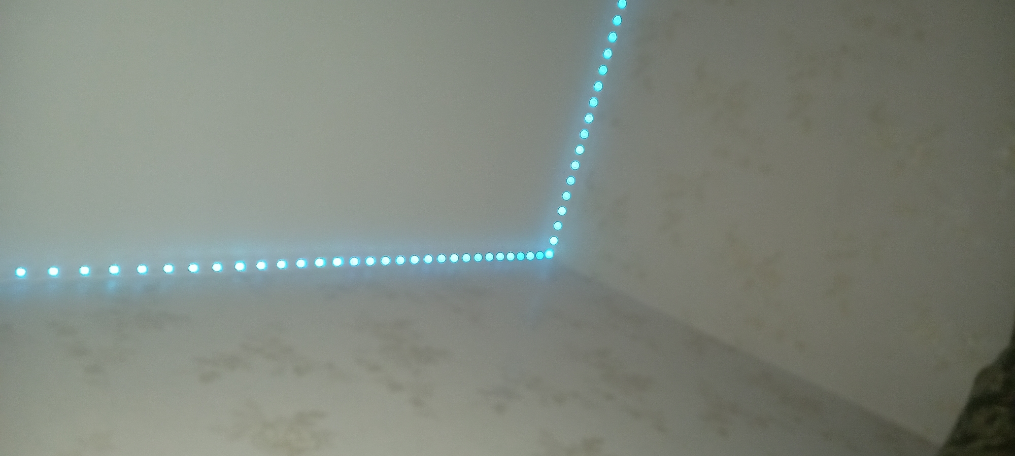 An interior shot shows a corner where two walls meet, forming a right angle. Along the top edges of these walls, a string of blue LED lights follows the contour of the corner. The lights are evenly spaced and emit a soft, consistent glow. The walls themselves are painted a light color and have a subtle, faded wallpaper pattern with what appears to be floral or abstract motifs. The lighting is dim, with the blue LEDs being the primary light source, casting a faint illumination on the surrounding surfaces. The perspective suggests the photo was taken from a low angle, looking upwards into the corner. The specific location in Netishyn, Ukraine, cannot be determined from the visual information present in the image.