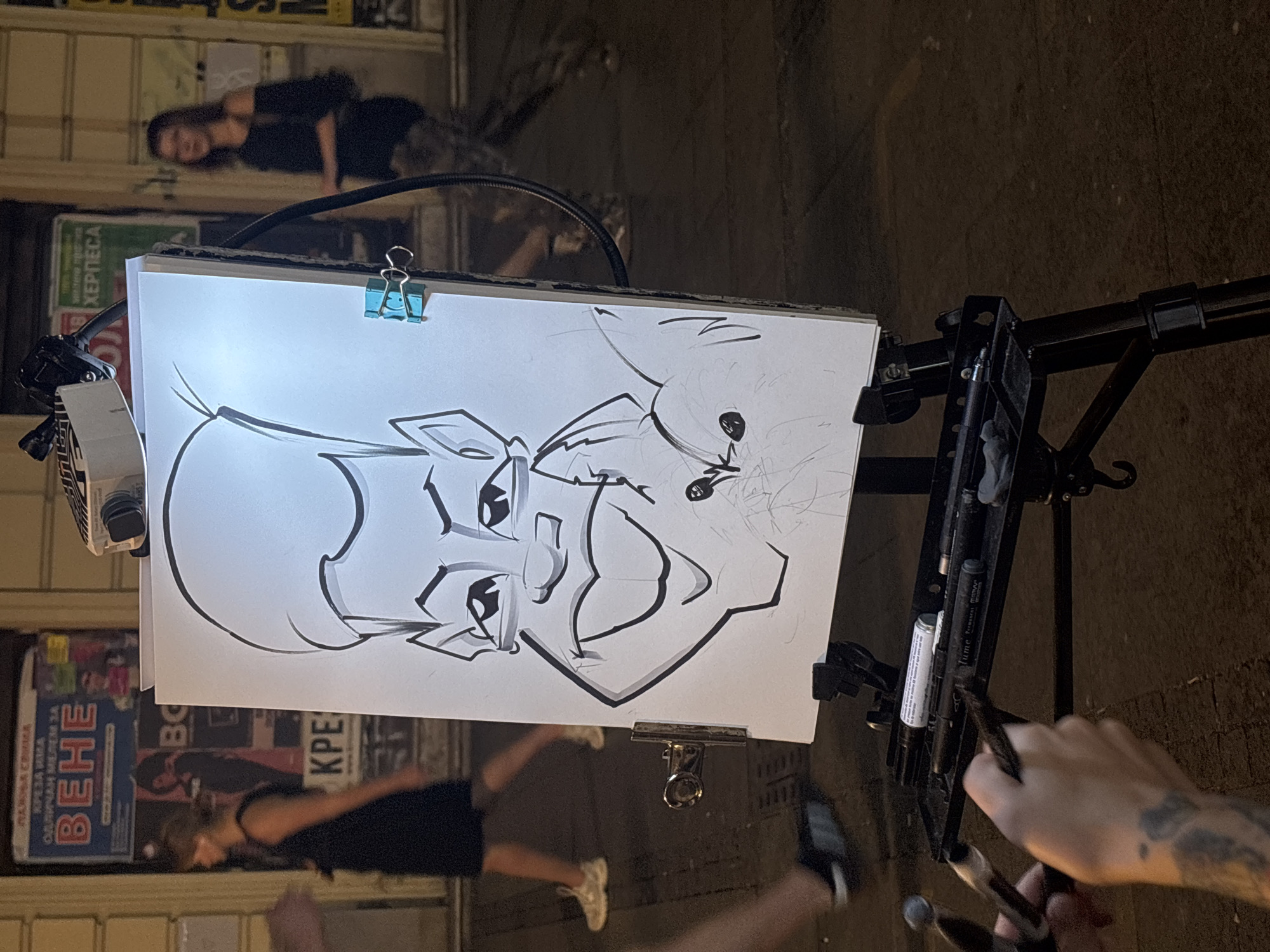 In Belgrade, Serbia, an outdoor artist is captured at night working on a caricature of a person. The caricature features a man with a large head, spiky hair, and exaggerated facial features, including a wide smile and prominent eyebrows. The drawing is illuminated by a bright light source positioned behind the canvas, casting a glow on the artwork and the artist's supplies.
The artist's hands are visible in the foreground, holding drawing tools. A tray on the easel holds several markers and pens, ready for use. In the background, pedestrians walk by, their forms blurred due to the low light conditions and movement. A woman in a dark dress and sneakers walks past on the left, while another woman in a black top and patterned pants is visible on the right.
Behind the easel, posters are affixed to a building facade. One poster features bold red text that reads "ВЕНЕ" (Vene), possibly indicating an event or performance. Another poster has green text that reads "ХЕРПЕС" (Herpes). The overall setting appears to be a street or pedestrian area, with cobblestone or paved ground visible. The scene is dynamic, with the artist engaged in their work amidst the flow of city life at night.
