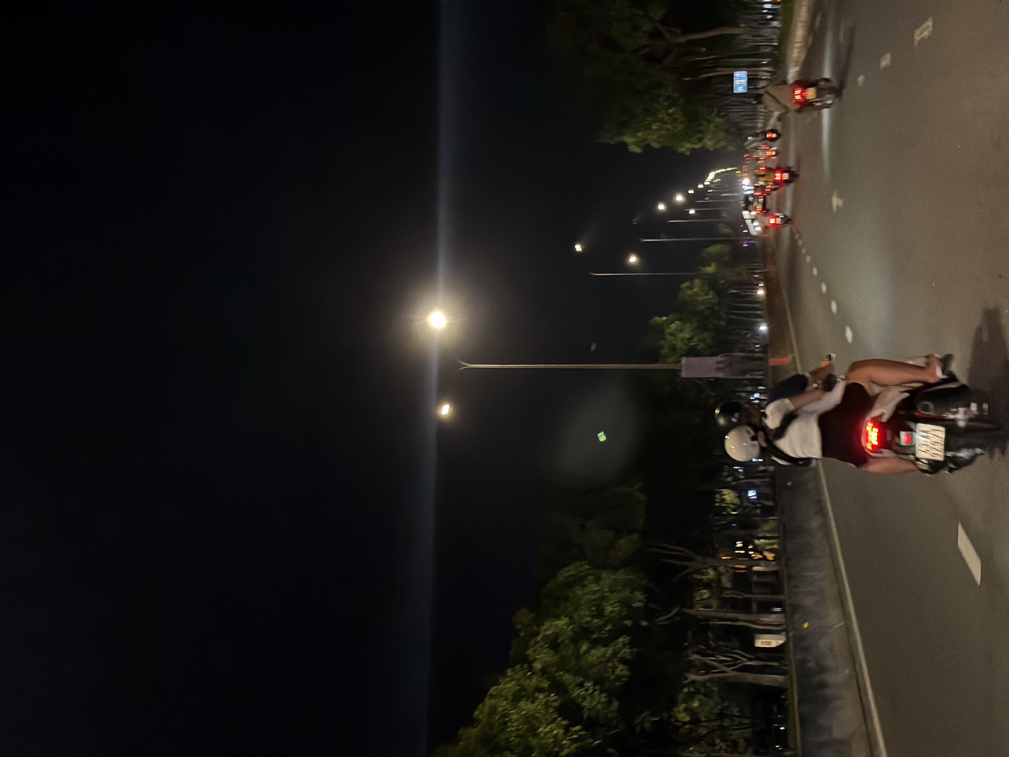 This is a nighttime, eye-level, medium shot of a busy road in Da Nang, Vietnam, possibly along the Han River embankment given the presence of trees lining the route and the general urban feel. The foreground is dominated by a motorcycle carrying two people. The rider, wearing a dark helmet, is driving, while the passenger, a female with a white helmet, is seated behind, embracing the rider. The motorcycle's license plate is visible, displaying the characters "92A1 075.77". The road is paved and marked with white dashed lines. Several other motorcycles, some with passengers, are traveling in the same direction ahead, their red taillights creating streaks of light in the darkness. On the left side of the road, dense foliage from trees forms a dark, green wall, interspersed with streetlights. On the right, more trees are visible, with further streetlights illuminating the road. The sky is dark, indicating it's late at night, and a bright street lamp creates a lens flare effect across the upper portion of the image. The overall atmosphere is one of a lively urban night scene, with multiple vehicles navigating the illuminated thoroughfare.