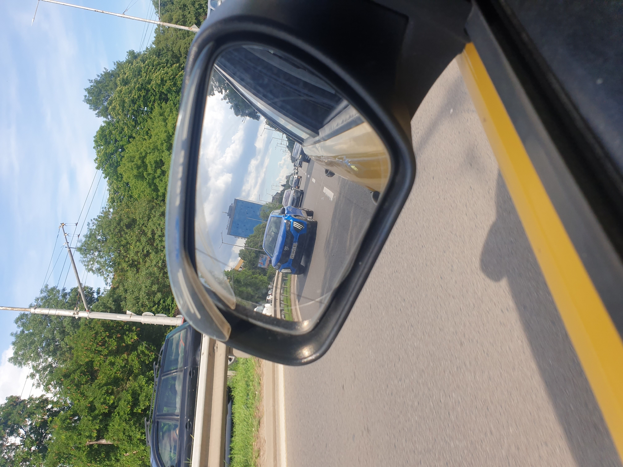 The image captures a view from a vehicle in traffic on a sunny day in Sofia, Bulgaria. A black station wagon is visible in the foreground on the left. In the side mirror of the vehicle, a blue car with a license plate is seen on the road. Further in the reflection, other cars are driving in traffic, and a tall blue building is visible in the distance. The road is paved, with painted lane markings, and lined with trees. Overhead power lines are also present. The overall scene depicts a typical urban traffic situation.