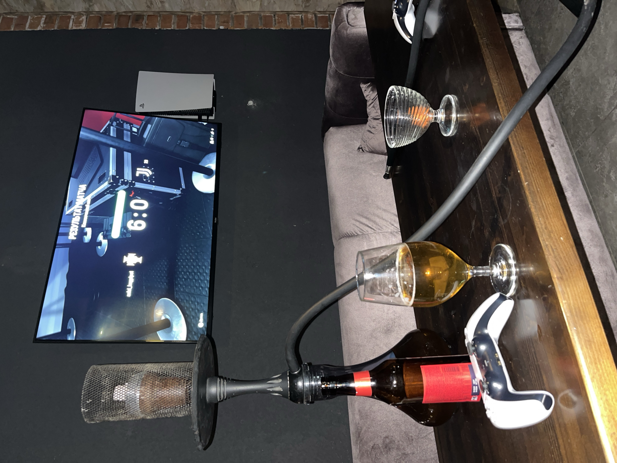 This indoor shot, taken in Akulovo (Gp Kubinka), Russia, depicts a leisure setup with gaming, hookah, and drinks. The scene is illuminated by artificial light, suggesting evening or nighttime.
On the left, a large flat-screen TV is mounted on a dark wall, displaying a video game, likely a football (soccer) match, given the "6:0" score and the partially visible "J" logo, possibly representing Juventus. Above the TV, a white PlayStation 5 console stands vertically on a shelf. Below the TV, a hookah is positioned on the floor, with its metal mesh cover visible, containing what appears to be shisha tobacco.
To the right, a dark wooden table holds various items. A hookah hose extends from the floor-standing base to the table, connecting to an empty amber-colored bottle, which serves as the hookah's water base. Next to the bottle, a white PlayStation 5 DualSense controller rests on the table. Two glasses are also present; one is an empty, ribbed clear glass, and the other, a wine glass, contains a yellow-amber liquid, likely a beverage like beer or white wine.
Behind the table, a dark grey upholstered sofa or couch is partially visible. The wall above the TV is dark, with a strip of exposed reddish-brown brickwork along the top edge, suggesting an industrial or rustic design element. The visible text on the TV screen includes "6:0" and "РЕЗУЛЬТАТ МАТЧА" (Match Result), confirming the Russian language context. A small "Haier" logo is also discernible on the bottom right of the TV screen. The PlayStation "PS" logo is visible on the console and controller.