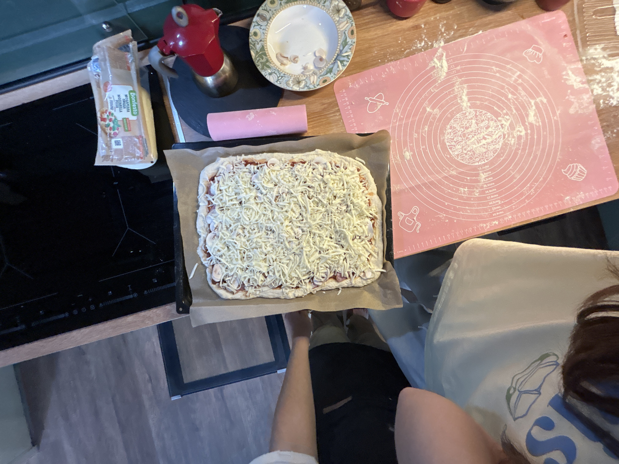 The image depicts a kitchen scene, likely in Shepelyovo, Russia, where a rectangular pizza is being prepared. The pizza, topped with a generous amount of shredded cheese, rests on a baking sheet lined with parchment paper. In the background, a stovetop is visible, and to the right, a red moka pot and a decorative plate with a floral pattern are present. A pink silicone baking mat, dusted with flour and featuring circular guides and icons of kitchen utensils, is prominently placed in the foreground. A person's arm and shoulder are visible on the left side of the frame, suggesting their involvement in the pizza-making process. Another person's head and hair are visible at the bottom of the image, looking down at the baking mat. The overall environment appears to be a home kitchen with wooden flooring and countertops.