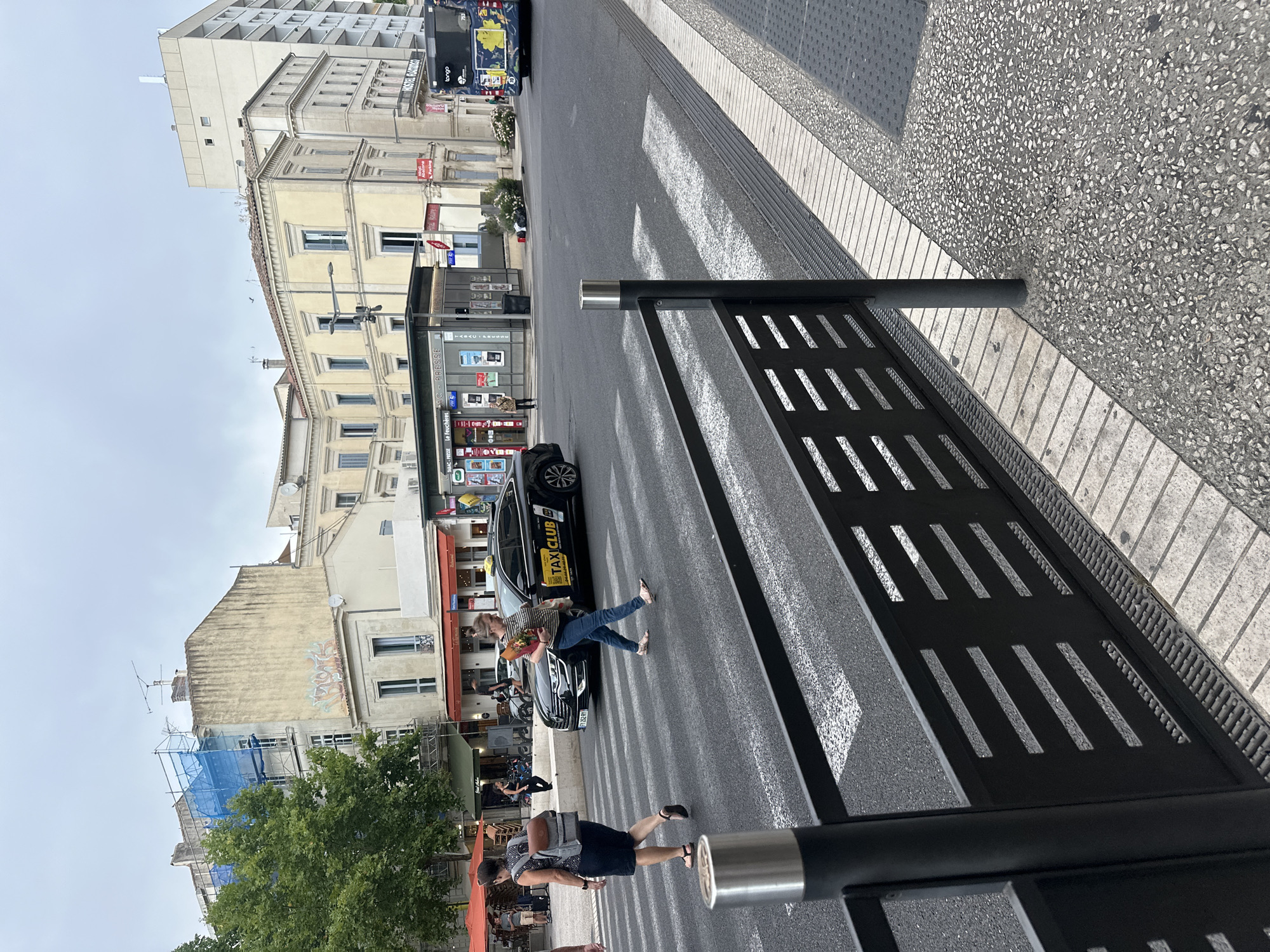 In Nîmes, France, at the intersection of Avenue Feuchères and Boulevard Victor Hugo, a pedestrian crossing is depicted. Two women are crossing the street. The woman on the left is wearing a dark blue dress, a backpack, and sandals. The woman on the right is wearing jeans, a striped shirt, and sandals, carrying a bouquet of flowers. A black taxi with "TAXICLUB" signage is stopped at the pedestrian crossing. In the background, several buildings line the street, including a multi-story building with scaffolding, a yellow facade building, and a bus with a floral design. A bus stop kiosk is also visible. The sky is overcast.
