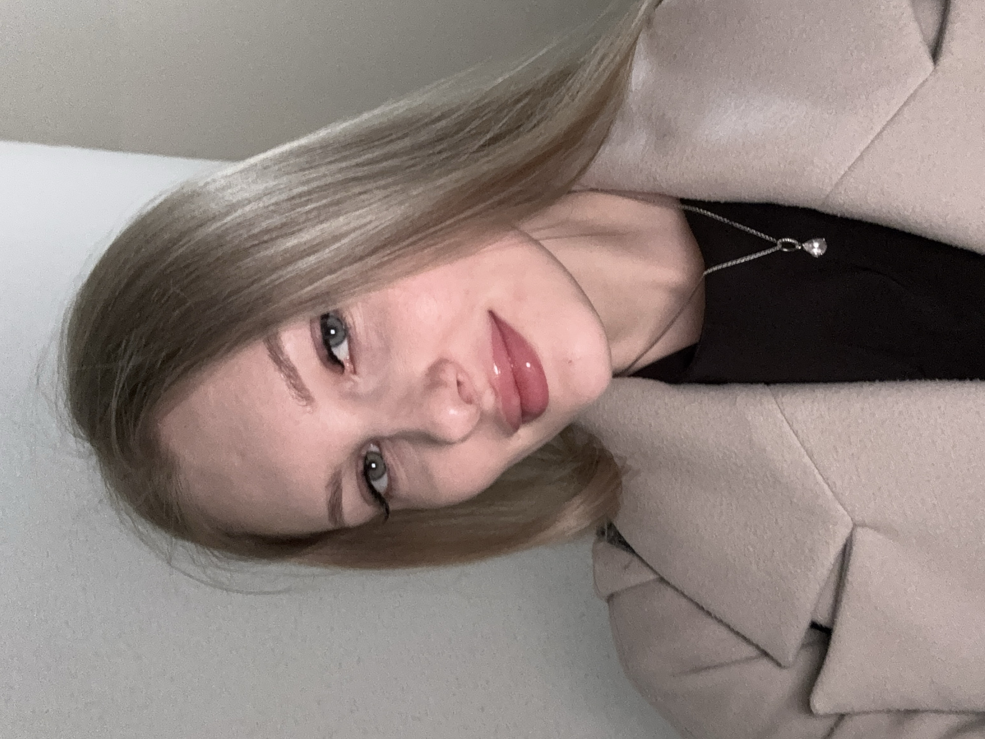 A young woman with light blonde hair and pale skin poses for a close-up, indoor portrait. Her eyes are light-colored and she has subtle makeup. She is wearing a black top underneath a light beige jacket with a distinct collar. A delicate silver necklace with a teardrop-shaped pendant is visible around her neck. The background appears to be a plain, light-colored wall. There is no information in the image to indicate the location. The information of Perm, Russia is not discernible from this image.