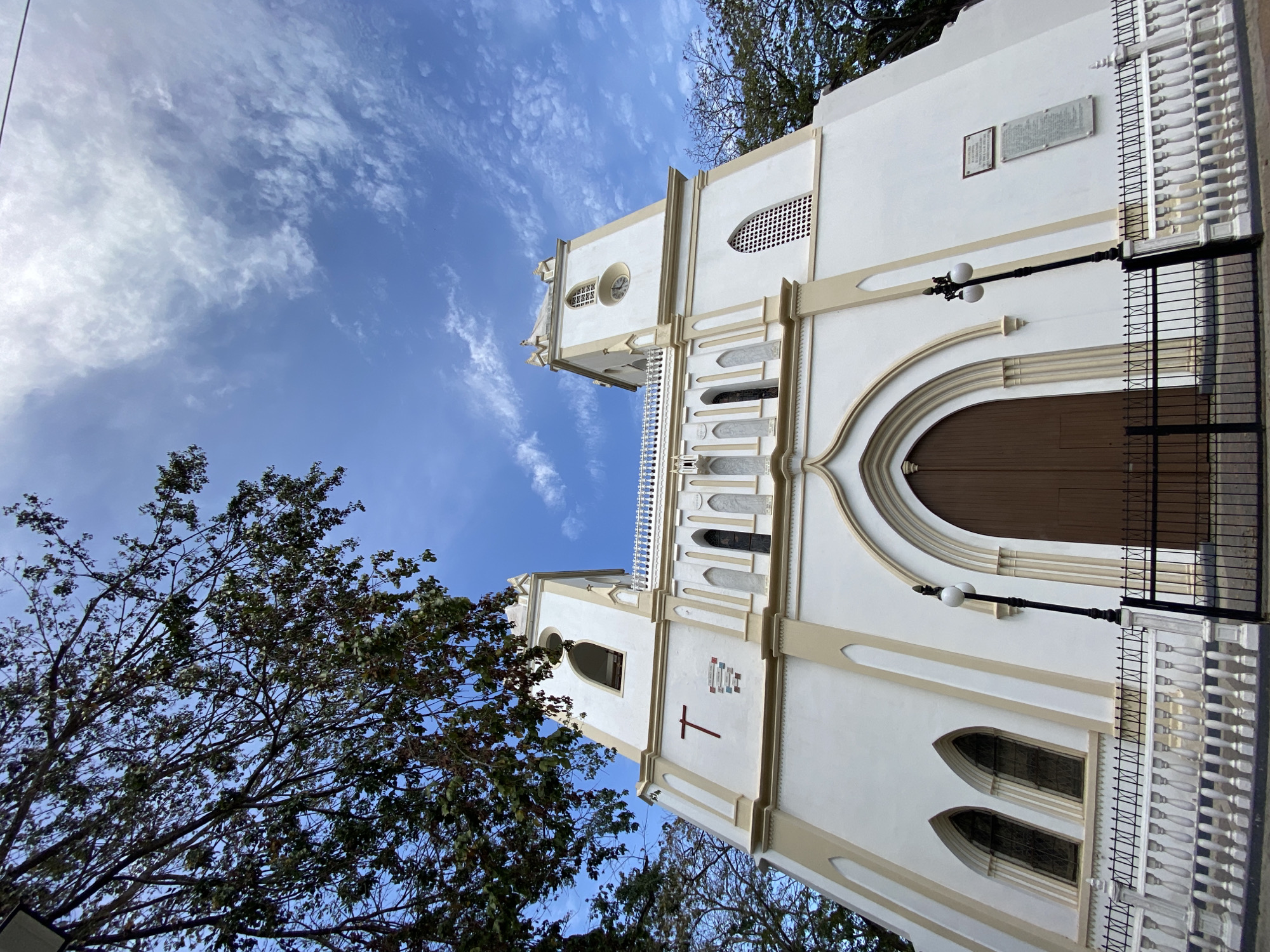 The image shows the Iglesia de San Nicolás de Bari, a church located in Porlamar, Venezuela. The church is white with beige trim and has two towers. A red cross is visible on one of the towers. A large brown wooden door serves as the main entrance. There are ornate windows and balustrades. Trees with green leaves are visible on the left and bottom of the image, partially obscuring the view of the church. The sky is blue with white, wispy clouds. A black wrought iron fence runs along the right side of the church. A lamppost with two white globes is attached to the building near the entrance.