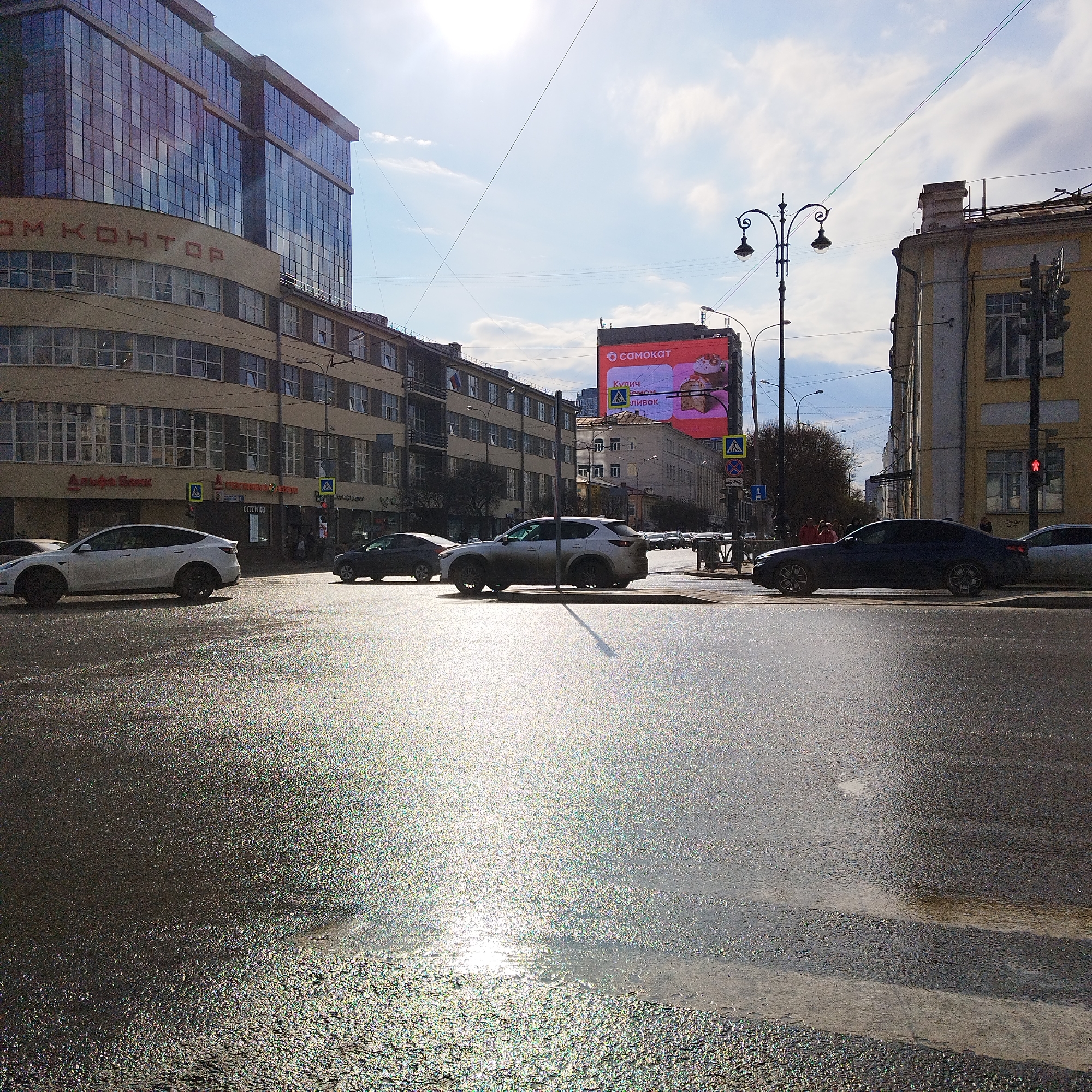 The image captures an urban street scene, likely an intersection, under bright daylight. The sun is low in the sky, causing strong specular reflections on the wet asphalt surface in the foreground and midground, suggesting either recent rain or a persistent wet patch.
On the left, a large, multi-story building with a modern glass facade rises above an older, curved structure. The older part of this building features the visible text "ДОМ КОНТОР" (House of Offices/Bureaus) and "Альфа Банк" (Alfa Bank) on its lower levels, with "ОПТИКА" (Optics) further down the street. Across the intersection, on the right, stands another multi-story building with a yellow facade.
Several vehicles are present on the road, including a white Tesla Model Y in the foreground, a gray SUV further back, and a dark sedan closer to the right. Cars appear to be in motion or stopped at the intersection. A few indistinct human figures are visible on the sidewalks in the distance.
In the midground, a tall, ornate street lamp stands prominently. Above the buildings on the right, a large digital billboard displays an advertisement for "самокат" (Samokat, a delivery service) and promotes "Кулич СМОМ" (Kulich, likely an Easter cake/bread) with "СЛИВОК" (cream/creamy) also visible, indicating a Russian-speaking location. Pedestrian crossing signs are also visible, and a traffic light on the right displays a red signal. The sky is bright with some scattered clouds.