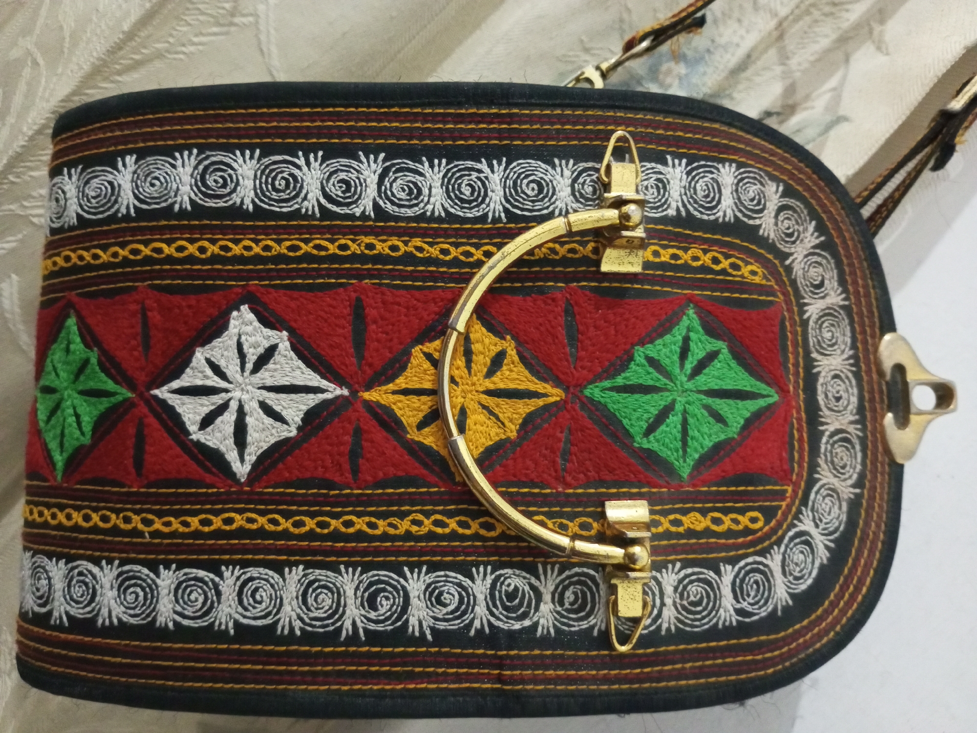 A close-up shows a black bag with embroidered patterns of white spirals, yellow chains, and large diamond shapes filled with white, yellow, and green starbursts against a red background. The bag features a brass handle and clasps, indicating its closure mechanism. The bag appears to be resting on a textured light-colored surface, with a curtain or fabric visible in the background. The image was likely taken indoors, given the lighting and surroundings. The location in the city of Lhokseumawe, Indonesia cannot be determined from the image alone.