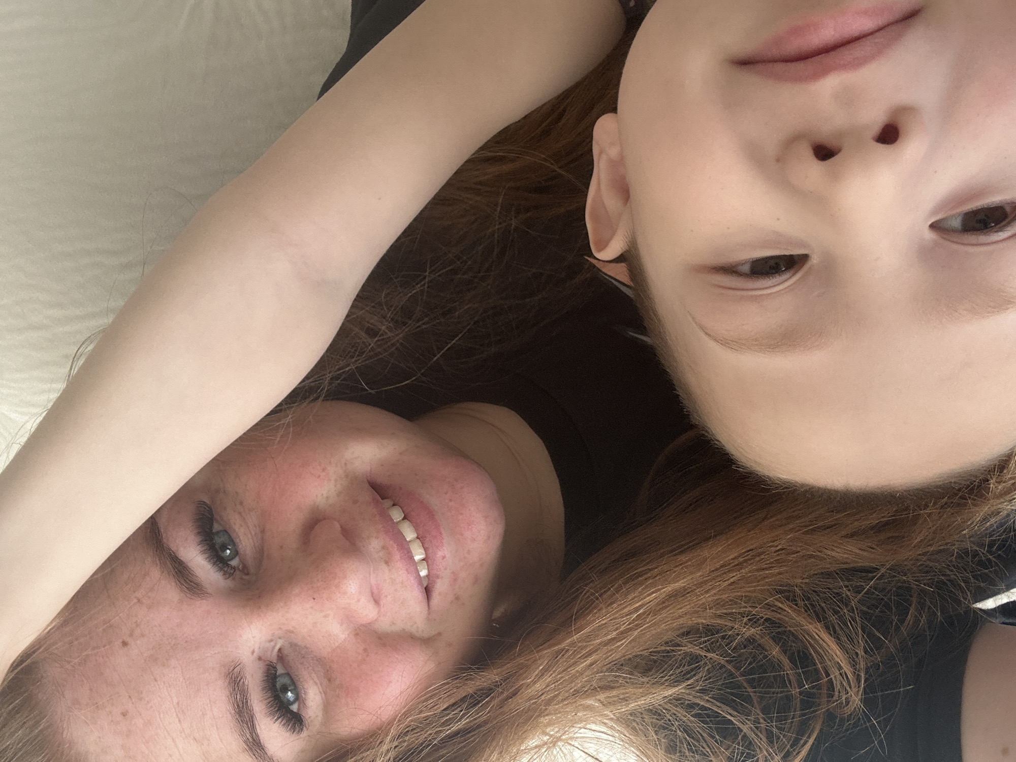 Two people, an adult and a child, are lying down close together, facing the camera at an upside-down angle. The adult has fair skin with freckles, blue eyes, and red hair, and is smiling. The child has fair skin, light brown eyes, and lighter brown hair. Both are wearing dark clothing. The background appears to be a light-colored surface, possibly a bed or couch. There is no information to determine the city of Chaikovsky, Russia.