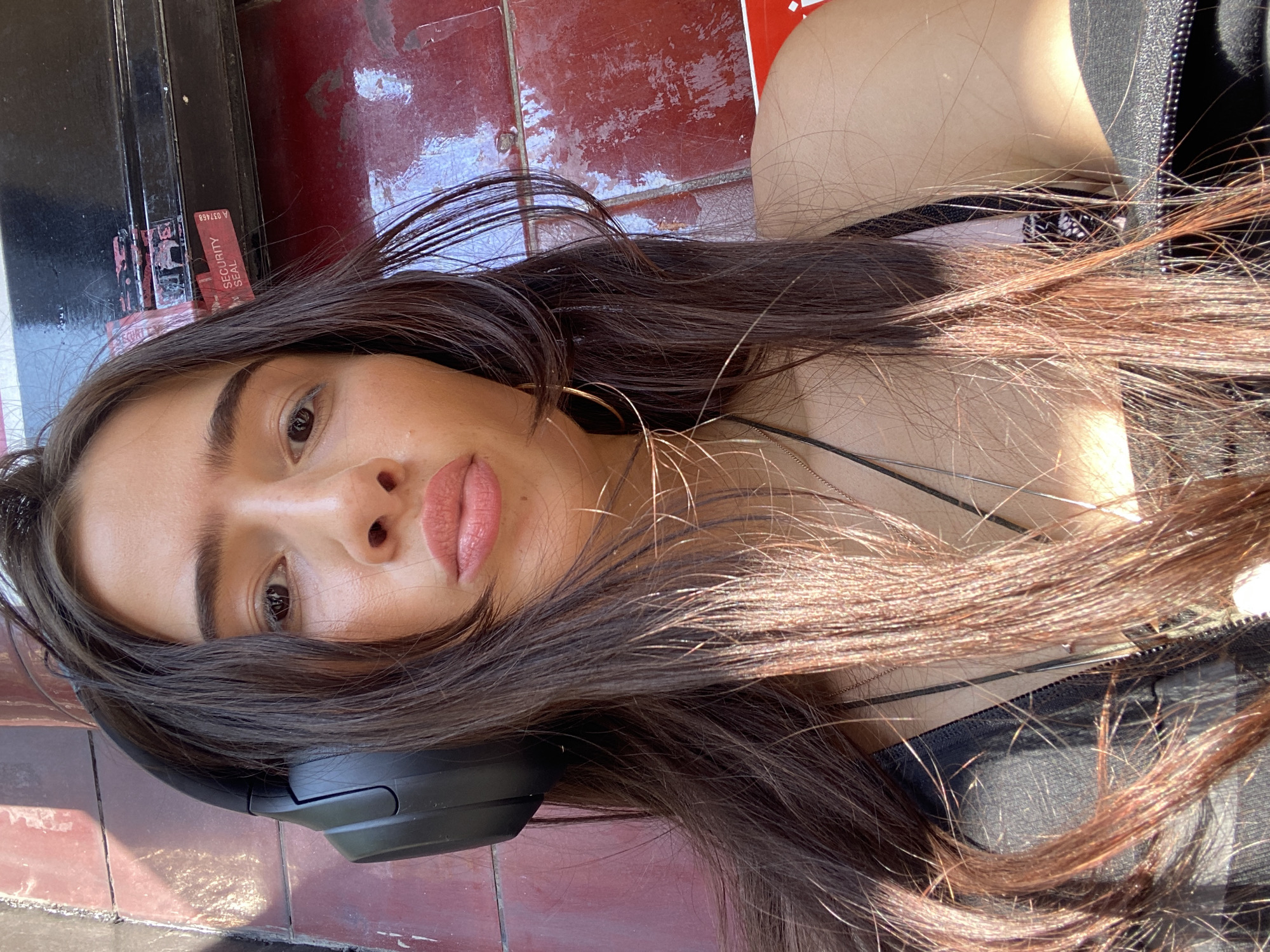 A young woman lies on a surface that appears to be part of a building in London, United Kingdom. Her head rests on dark-colored headphones. She has long, dark brown hair, fair skin, and full lips. She is wearing a black top with a zipper. A thin gold hoop earring is visible. The background includes a wall with a red and black tiled pattern, and a sticker with "SECURITY SEAL" printed on it. The lighting is bright, suggesting a sunny day. The overall setting suggests an urban environment.