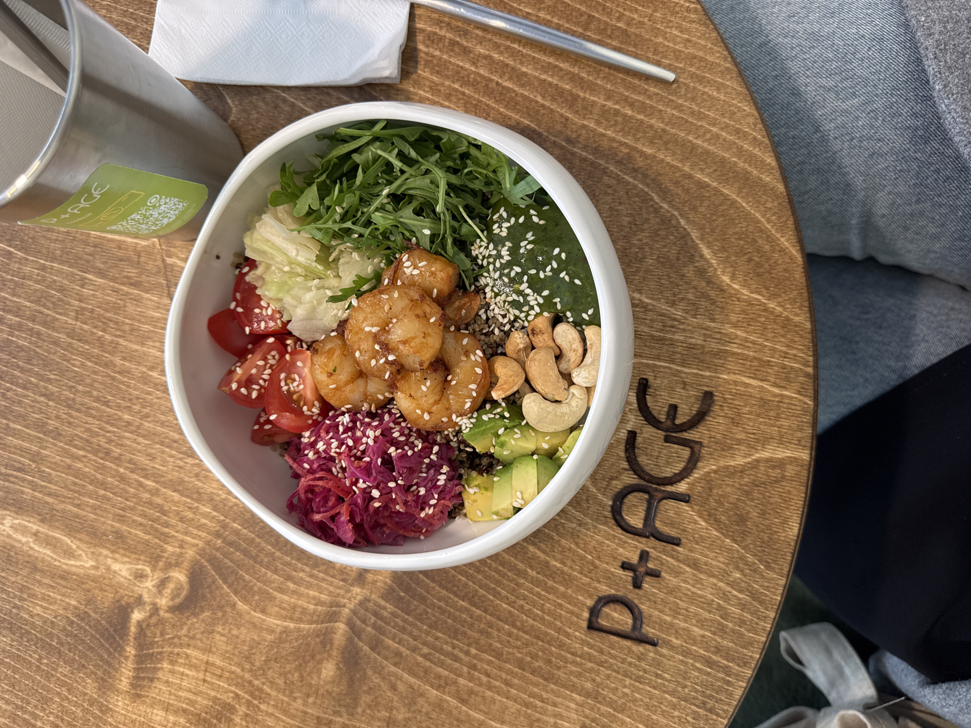 A top-down shot captures a colorful salad bowl, a beverage container, a napkin, and utensils on a wood-grained surface, seemingly a table. A person in denim jeans is seated at the table.
The salad bowl, a focal point, is filled with a variety of ingredients: sliced tomatoes, lettuce, arugula, cooked shrimp with sesame seeds, shredded red cabbage, sliced avocado, quinoa, toasted cashews, and pickled cucumber with sesame seeds.
The beverage container is partially visible on the left, along with a green label containing the letters "PACE" and a QR code. A white napkin and a utensil (possibly a spoon or fork) are placed on the upper left.
On the table, the word "P+ACE" is engraved, seemingly a branding detail of the location. The table itself has a distinct circular shape.
Although the scene is set indoors, and based on the "P+ACE" detail and a Google search, it is probable that this photograph was taken in a restaurant in Sochi, Russia.