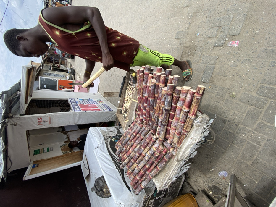 The image depicts an outdoor street scene in Ado, Nigeria. In the foreground and midground, a young Black male stands on the right, positioned in profile facing left. He wears a dark reddish-brown sleeveless top with yellow star patterns, light green track pants with black stripes, and patterned slides. He is actively peeling a segment of sugarcane with a silver-bladed knife, holding the cane with his left hand. Directly in front of him, a large quantity of reddish-brown sugarcane stalks, some partially peeled, are arranged in rows on a makeshift white-covered stall or cart. To the right of the sugarcane display, on the same stall, are piles of sugarcane peelings. Behind this setup, a small, white-paneled kiosk with a corrugated metal roof is visible, bearing "POS" and "NANA VENTURE" signage. Inside the kiosk, a second Black male is seated. The ground is paved with grey interlocking bricks, showing minor debris. Other distant structures, likely market stalls, and a vehicle are visible in the background under an overcast sky.