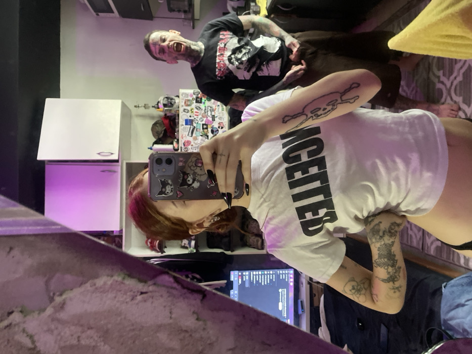 An indoor mirror selfie captured in Buenos Aires, Argentina, depicts two individuals within a residential setting. In the foreground, a female subject with reddish-brown hair, adorned with multiple ear piercings and arm tattoos, holds a dark smartphone featuring a "Kuromi" character case to photograph her reflection. She wears a white t-shirt displaying reversed black text ("DETTEGCM") and black underwear. Her left arm exhibits floral tattoos, while her right forearm bears an outline tattoo of a stylized character. Behind her, to the right, a male subject with extensive facial, neck, and arm tattoos, including visible piercings, stands in brown shorts and a black graphic t-shirt. He presents an exaggerated expression with his mouth wide open, looking towards the mirror. The background shows various domestic elements: a bed next to a monitor displaying text, a patterned floor, white wall-mounted cabinets, a refrigerator or similar appliance covered in numerous stickers, and shelving units holding miscellaneous items, including shoes. A purple ambient light source illuminates the left side of the scene, and the mirror's surface appears worn or textured on its left edge.