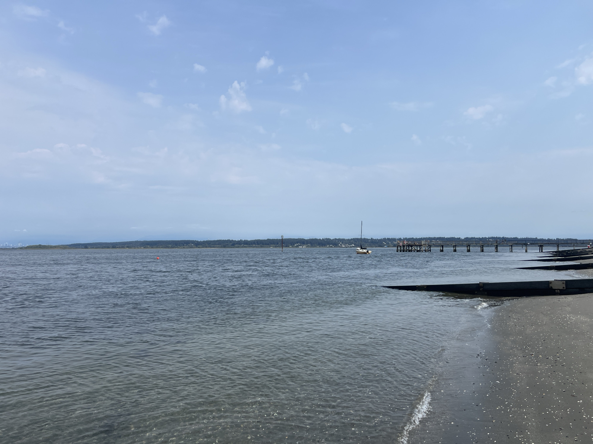 This is a wide shot of the ocean at the beach, likely in Surrey, Canada, under a partly cloudy sky. The water is a calm, dark blue, with gentle waves lapping at the dark sand. There are various structures along the shoreline, including a pier that extends out into the water and some dark, angled structures on the beach. A small sailboat can be seen in the distance. The horizon is lined with a distant treeline, and the city of Vancouver can be seen in the far distance.