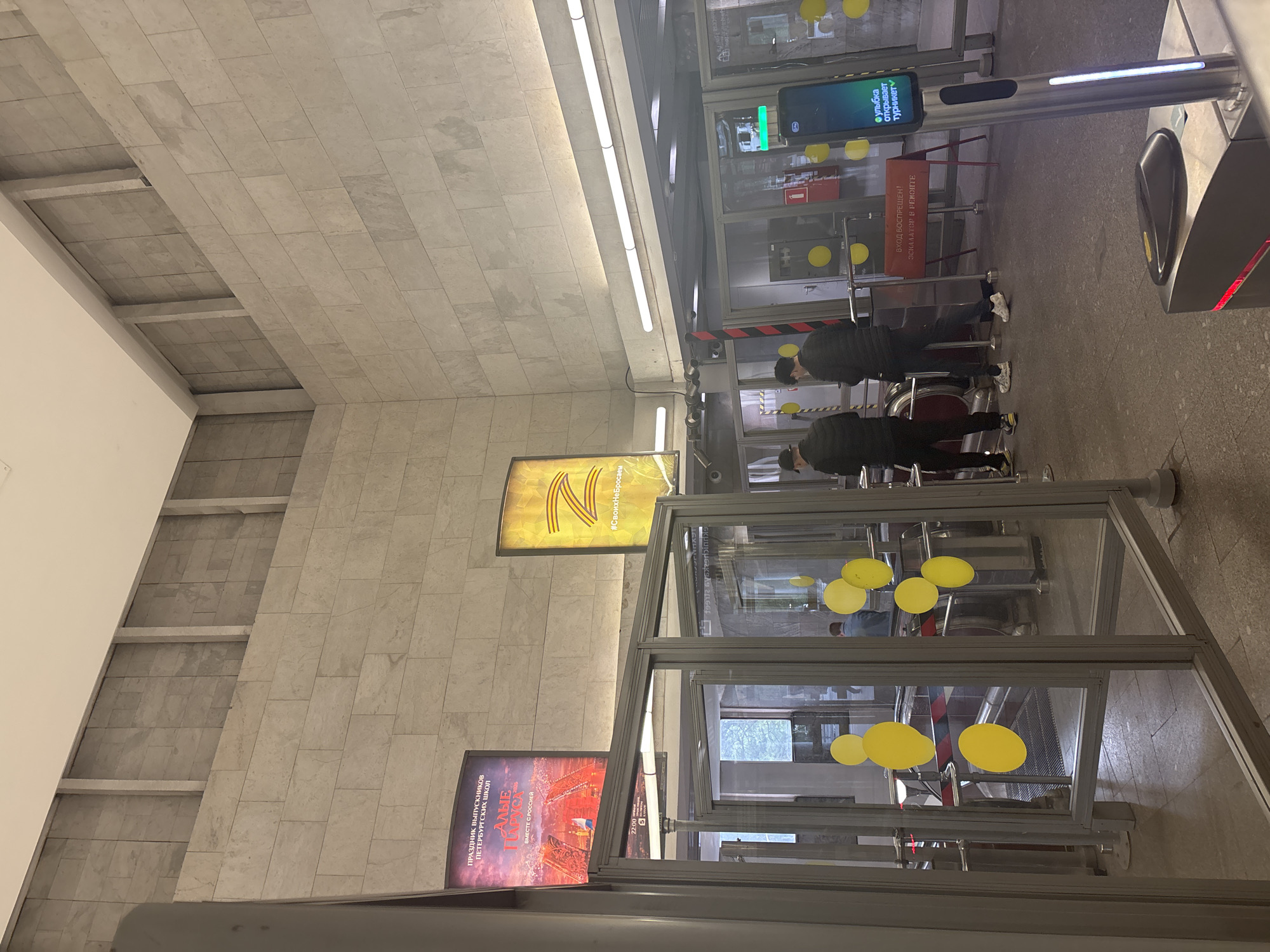 The image depicts an indoor scene, likely a subway station or public transit entrance in Sinyavino, Russia, given the context. The scene is characterized by modern architecture with tiled walls and fluorescent lighting. In the foreground, several glass-enclosed turnstiles are visible, with yellow circular decals on the glass. Two individuals are seen waiting near the turnstiles.
Prominently displayed are several posters. One poster, with a yellow background and a large "N" symbol, has the hashtag "#СвоихНеБросаем" (We don't abandon our own). Another poster, featuring a ship and fireworks, is titled "АЛЫЕ ПАРУСА" (Scarlet Sails) and advertises a "ПРАЗДНИК ВЫПУСКНИКОВ ПЕТЕРБУРГСКИХ школ" (Graduation Celebration for St. Petersburg Schools).
Further back, a red sign reads "ВХОД БОСПРЕЩЕН! ЭСКАЛАТОР В РЕМОНТЕ" (ENTRY FORBIDDEN! ESCALATOR UNDER REPAIR). A digital screen displays the text "улыбка открывает турникет" (A smile opens the turnstile). The overall atmosphere appears to be that of a public space with ongoing events or advertisements, possibly related to a celebration or a public service announcement. The lighting suggests it is not daytime outdoors, and the indoor setting implies controlled lighting conditions.