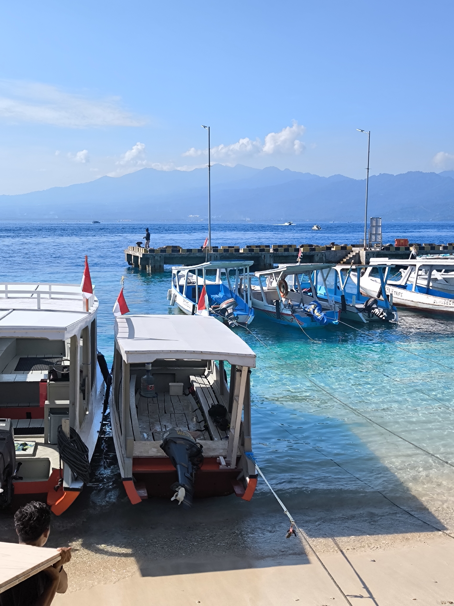 The image shows a waterfront scene in an unspecified location in Indonesia, presumably near a city denoted as Unknown. Several small boats are docked or moored at a pier or small harbor, with crystal-clear water revealing the sandy seabed.
To the left, two larger boats with white upper structures and red hulls are close to the shore, with at least one person visible near them. In the background, a pier extends into the water with individuals standing on it.
The boats docked at the pier are smaller, mostly blue and white, with what appear to be small passenger areas covered by canopies. The water around the pier is exceptionally clear, allowing a view of the underwater details.
In the distance, there are mountains under a clear blue sky dotted with a few clouds. Two light poles are also visible along the dock.