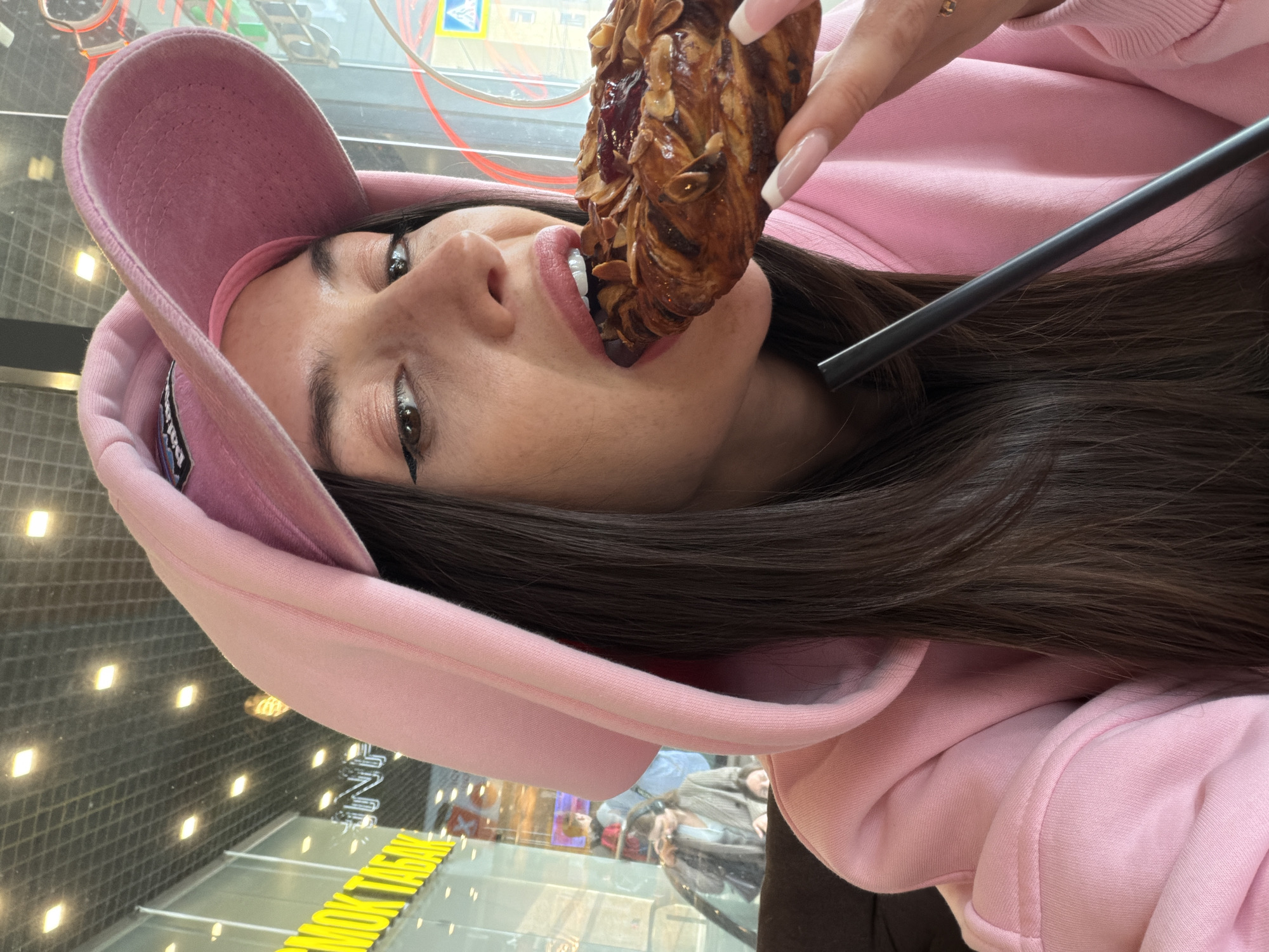 A person in a pink hoodie and cap is holding a pastry with almonds in front of their mouth. The pastry is slightly bitten into. A black straw is positioned near the person's cheek. The background shows a reflection of a street scene with yellow lettering. The reflection suggests the scene is taking place in Saint Petersburg, Russia.