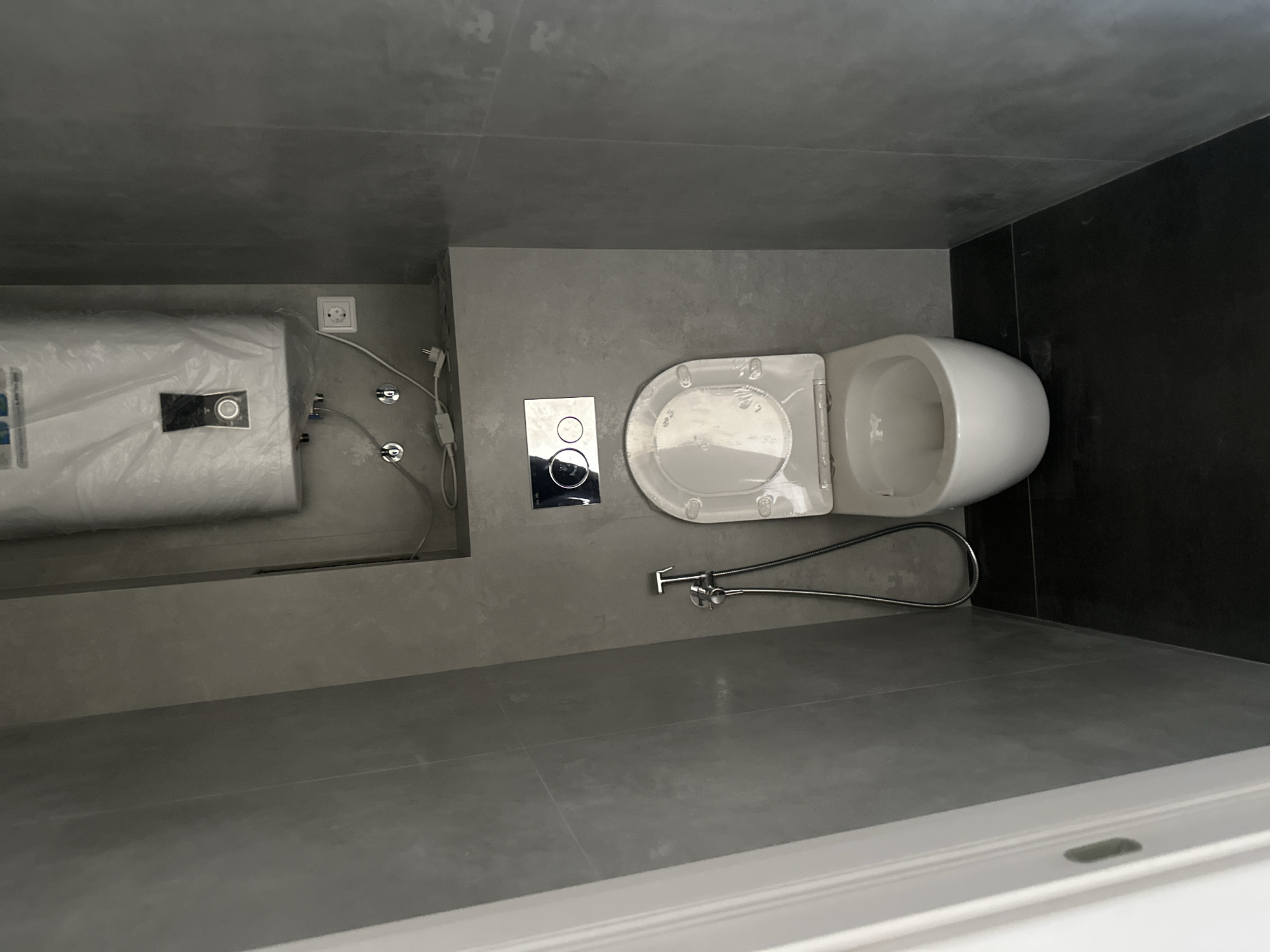 A modern bathroom in Novoaltaysk, Russia features a white toilet with a bidet spray attachment and a dual-flush toilet cistern actuator plate. Above the toilet, a white water heater is visible, covered in plastic. The walls are tiled with large grey slabs, and the floor is dark. The overall aesthetic is minimalist and contemporary.