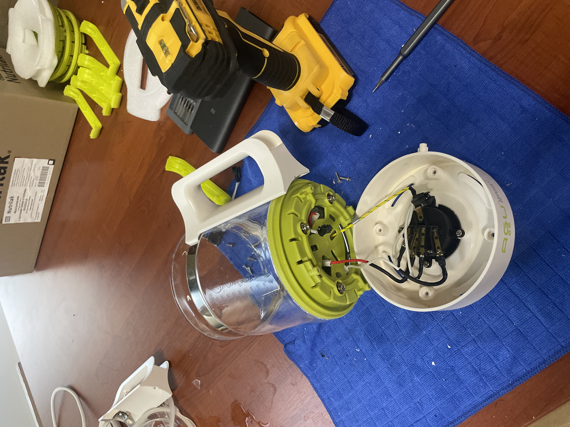 The image shows a disassembled blender on a wooden table. The blender's glass pitcher is detached from its base, which is also open, revealing wires and internal components. A yellow and black DeWalt drill and a small screwdriver are also present on the table, suggesting the blender is being repaired or disassembled. The background shows a cardboard box and some yellow plastic parts. The scene is well-lit and appears to be indoors. There are no people visible in the image. The city of Tbilisi, Georgia is not mentioned or indicated in the image.
