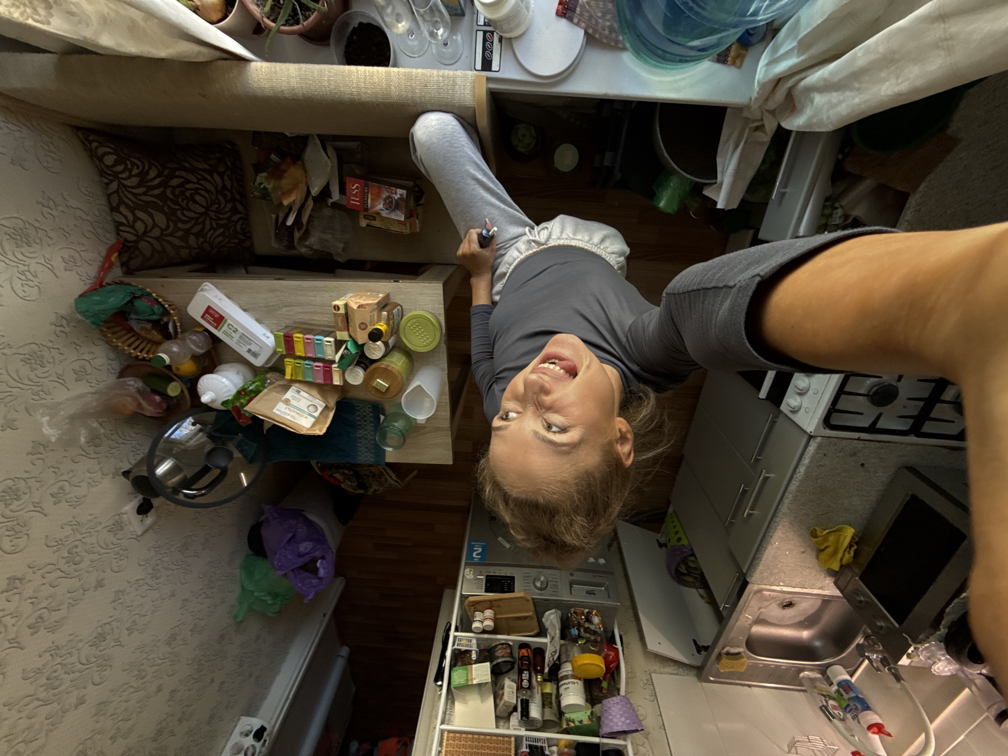 The image is a selfie taken from an overhead perspective, likely in an apartment in Chaikovsky, Russia. The subject is a light-skinned person with blonde hair, wearing a gray top and gray pants. The person is smiling at the camera and holding a small object in their hand.
The environment is a cluttered living space that combines kitchen and living areas. The floor is wooden and tiled. A cream colored wall with patterned wallpaper is visible. The sofa is beige, and there are various objects on the floor and the table: food, pot, and bottles. The kitchen includes a stove, oven, sink, and a rolling cart filled with various items like bottles, jars, and household supplies. A washing machine is also visible in the background. The image suggests a small and confined living space.