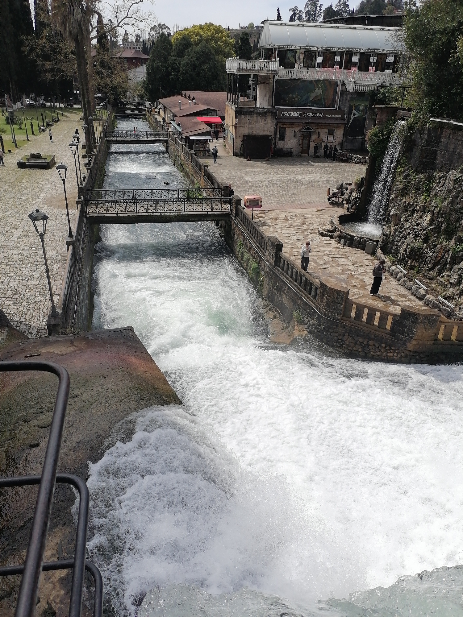 This elevated view captures an active urban landscape centered around a powerful, engineered waterway. In the immediate foreground, a torrent of intensely white, foamy water cascades downwards, indicating a significant drop or spillway, with a dark metal railing visible on the left.
The midground features a stone-lined canal with fast-flowing water, spanned by two ornate, dark metal pedestrian bridges. People are visible on the bridges and along the canal's banks. To the right of the canal, a paved area with patterned stonework hosts a small artificial waterfall cascading down a rock face. Two individuals stand on the path near this waterfall.
In the background, to the left, a wide cobblestone promenade is lined with old-fashioned street lamps and a mix of trees, including a prominent palm tree. Several people are walking along this path. To the right, a complex of buildings includes structures with red-tiled roofs and a larger building with a white, translucent arched roof. Large banners or murals decorate the facade of the larger building.
Visible text on one of the buildings reads "АБХАЗСКАЯ КОСМЕТИКА" (Abkhazian Cosmetics) and "Аква вита" (Aqua Vita). The scene is bathed in daylight under what appears to be an overcast or hazy sky, suggesting mild weather conditions. The overall impression is of a lively public space, possibly a park or a tourist attraction, where the powerful flow of water is a central and dynamic element.
