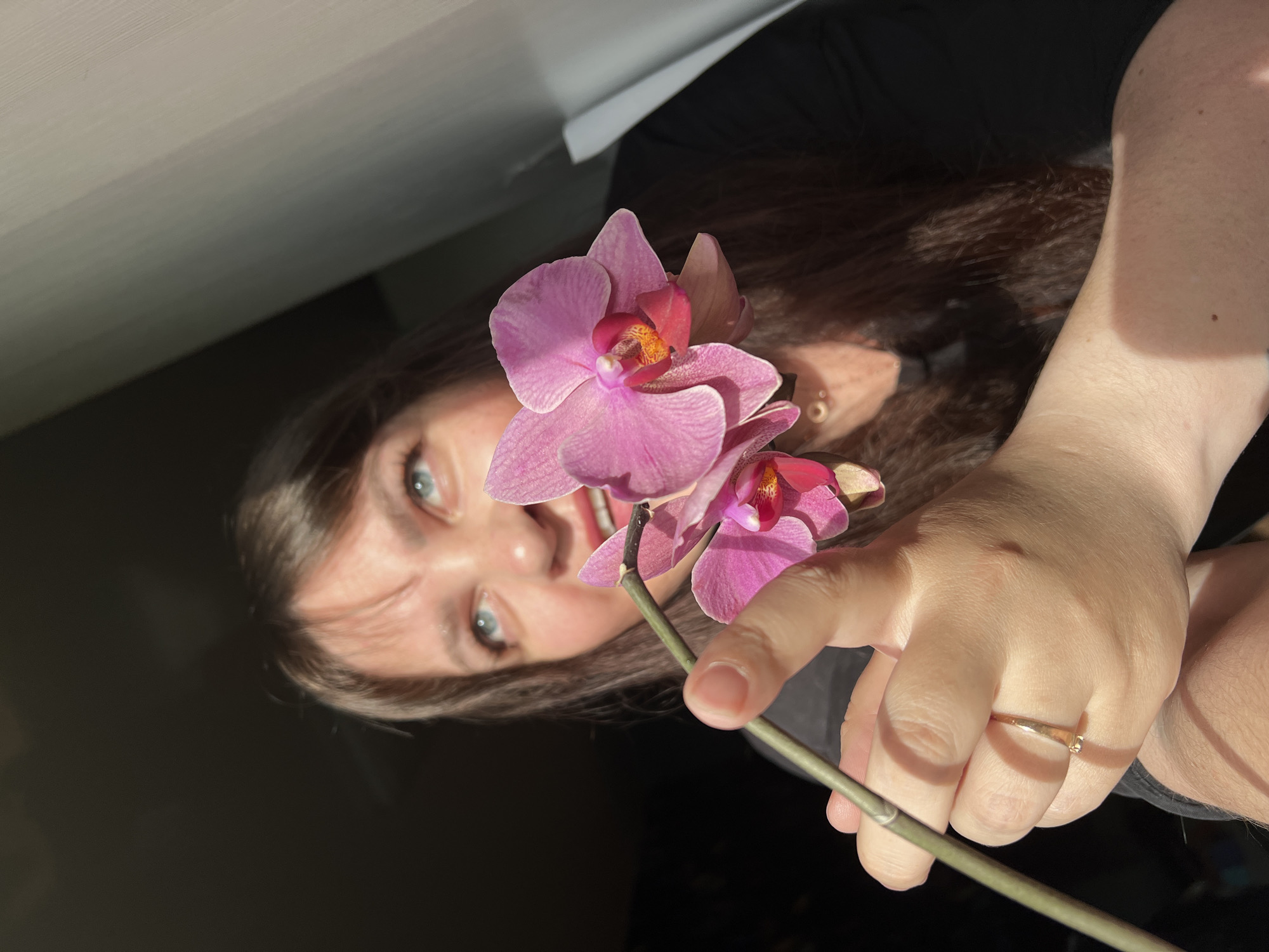 This image, taken indoors in Noviy, Russia, presents a close-up, rotated portrait of a person interacting with a flowering orchid plant. The subject, a young adult with long, dark brown hair and bright blue eyes, looks directly at the camera with a gentle smile. They are wearing a dark-colored top. Their right hand, adorned with a thin golden ring on the ring finger, is visible holding the stem of a vibrant pink Phalaenopsis orchid. Two prominent pink flowers with distinctive orange-yellow centers are in full bloom, with a smaller flower or bud further down the stem.
Strong, natural light illuminates the scene from the side, casting soft shadows on the hand and highlighting the delicate texture of the orchid petals. The background is largely dark and out of focus, with a bright, light-colored ceiling or wall visible in the upper portion of the frame, suggesting the image was captured indoors, possibly near a window during daytime. The overall impression is one of warmth and appreciation for nature. No text is visible. The original image is oriented 90 degrees clockwise.