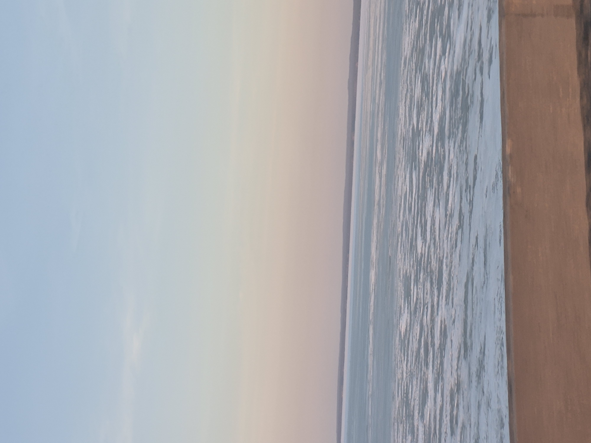 The image captures a view of the Oka River in Noviy, Russia. The water surface exhibits a textured appearance, potentially indicating ripples or small waves. The sky transitions from a pale blue hue on the left to a slightly warmer, pastel tone towards the center of the frame. A distant treeline is visible along the horizon. On the right side, there's a partial view of a concrete structure, possibly a bridge support or embankment.