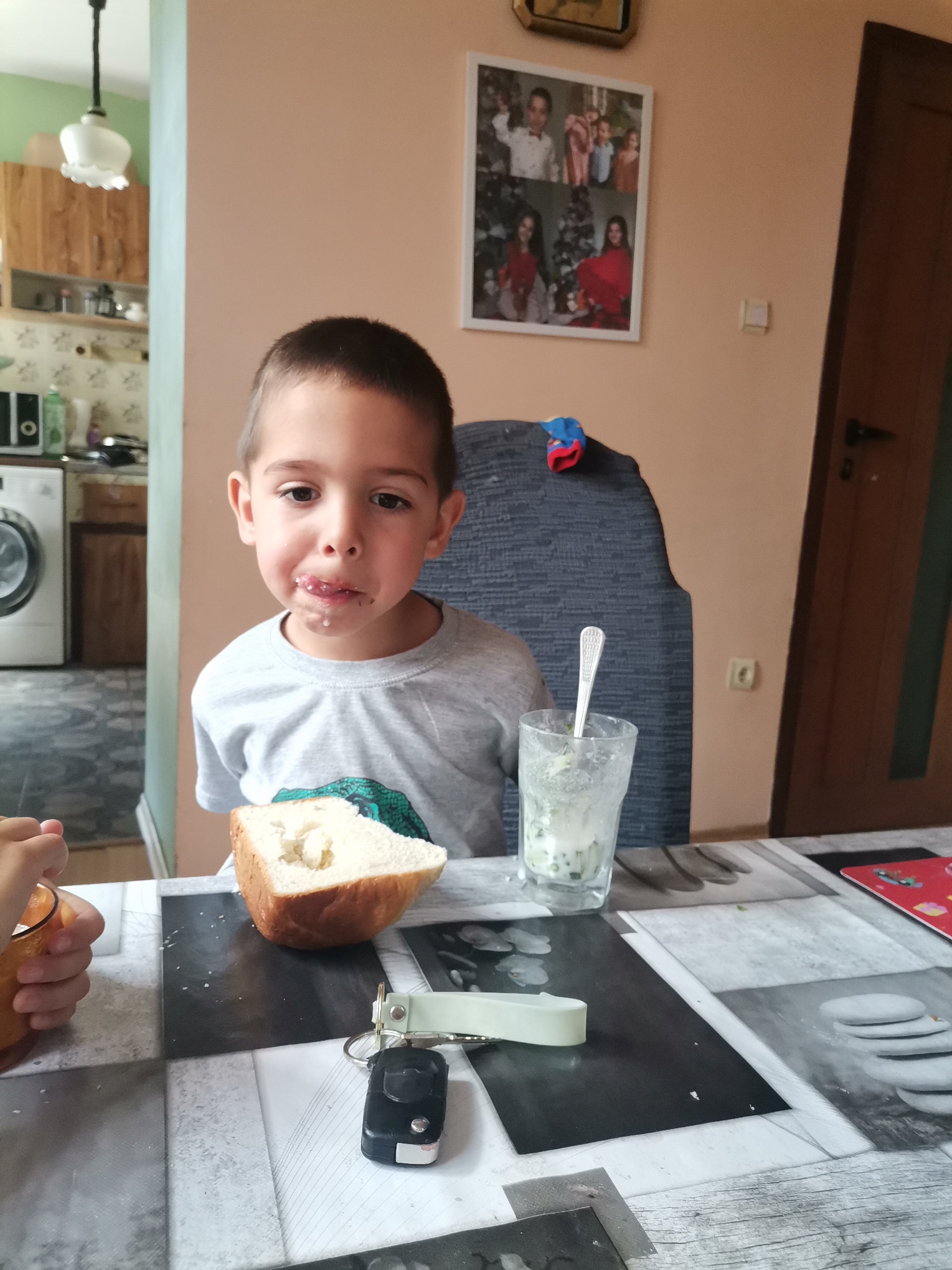A young boy sits at a table in a room with peach-colored walls. He has dark hair and a gray shirt. On the table are a glass with a spoon, a slice of bread, and a car key. A framed photo collage hangs on the wall behind him. A kitchen with a washing machine is visible in the background.
