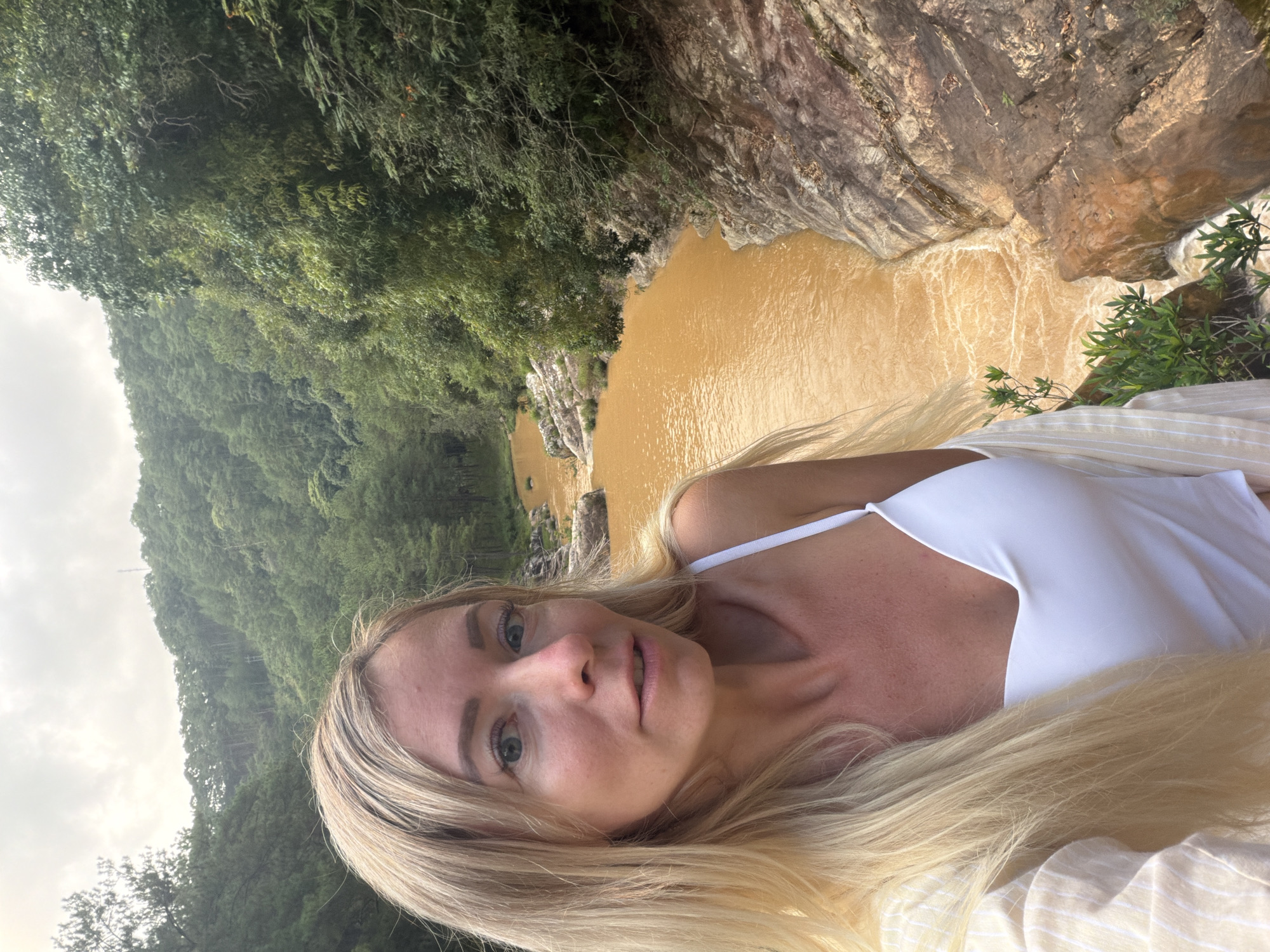 A woman with long blonde hair and a white top is taking a selfie by a muddy brown river flowing through a lush green forest in Păng Tiêng 1, Vietnam. The river is surrounded by rocky terrain and dense vegetation, with hills covered in trees in the background. The sky is overcast.