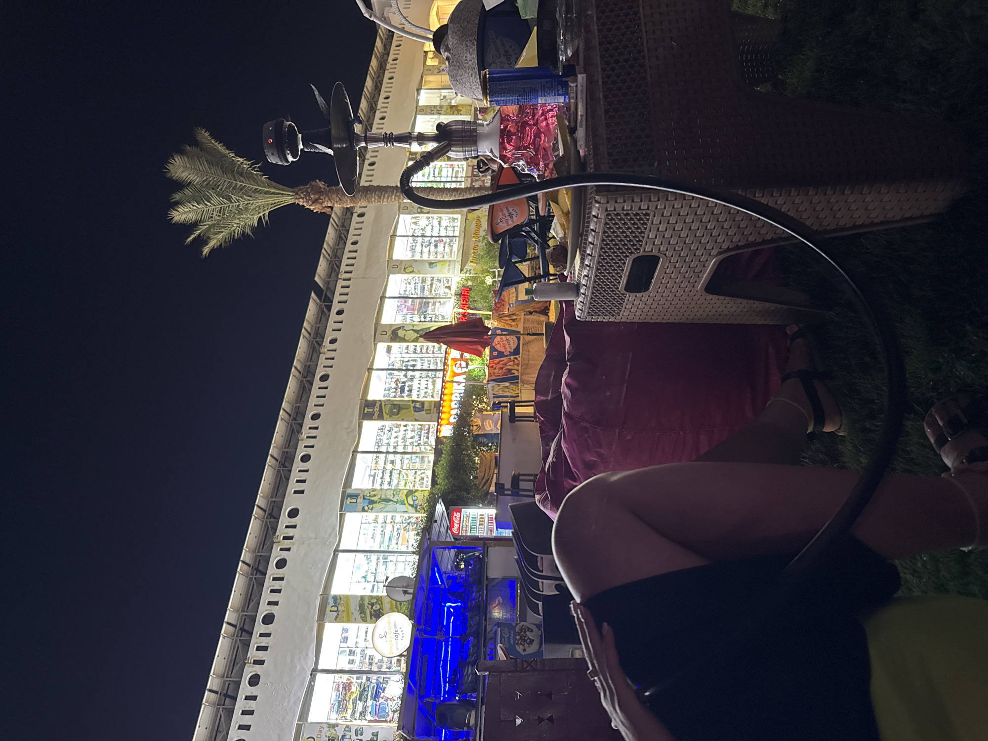 The image depicts an outdoor seating area at night, likely a cafe or lounge, in an unknown location in Egypt. In the foreground, a person's legs are visible, resting on a large, plush red beanbag chair. The person is wearing dark shorts and sandals. To the right of the beanbag is a woven wicker table, upon which sits a hookah pipe with a pink glass base. Other objects on the table include a plate with food, cigarettes, and a can. In the background, a building with large illuminated windows displays various merchandise. A palm tree stands in front of the building, silhouetted against the night sky. The overall atmosphere is relaxed and nocturnal, with artificial lighting illuminating the scene.
