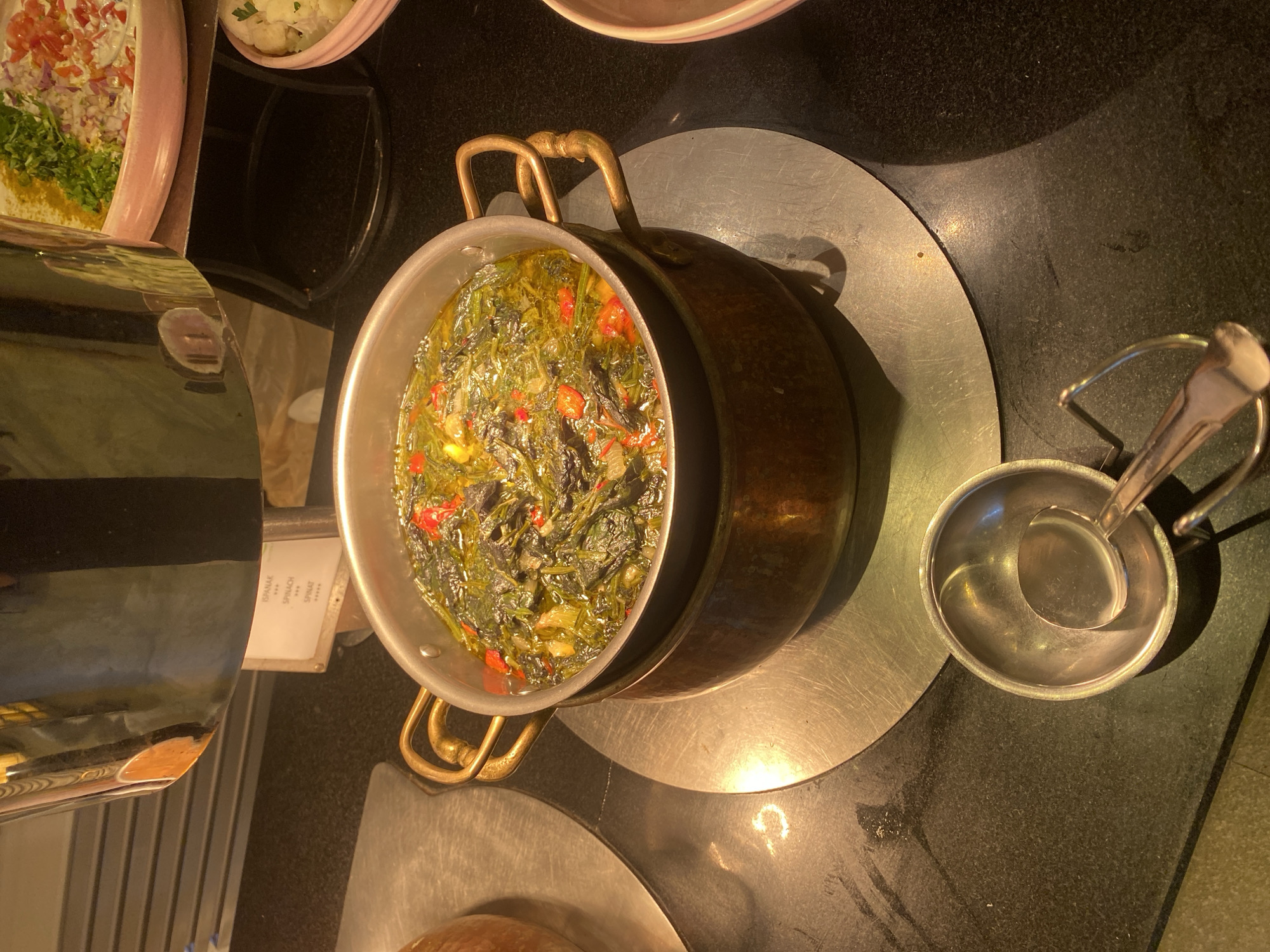 The image shows a close-up of a buffet serving dish at a food establishment in Aksu, Turkey. The dish contains a portion of cooked spinach with what appear to be red chili peppers in a greenish liquid, likely oil and broth. The serving dish is made of a shiny metal, possibly copper, with brass handles. It rests on a larger, circular metallic base. In the foreground, a silver serving ladle is placed inside a small silver bowl. Behind the serving dish, a metallic steam table unit is visible, and on the right side of the image, a pink serving bowl containing a salad-like mixture of chopped vegetables and herbs can be seen. A small white label is affixed to the steam table unit, with text that appears to include "ISPANAK", "SPINACH", and "SPICED". The overall environment suggests a self-service food setup, common in hotels or restaurants.