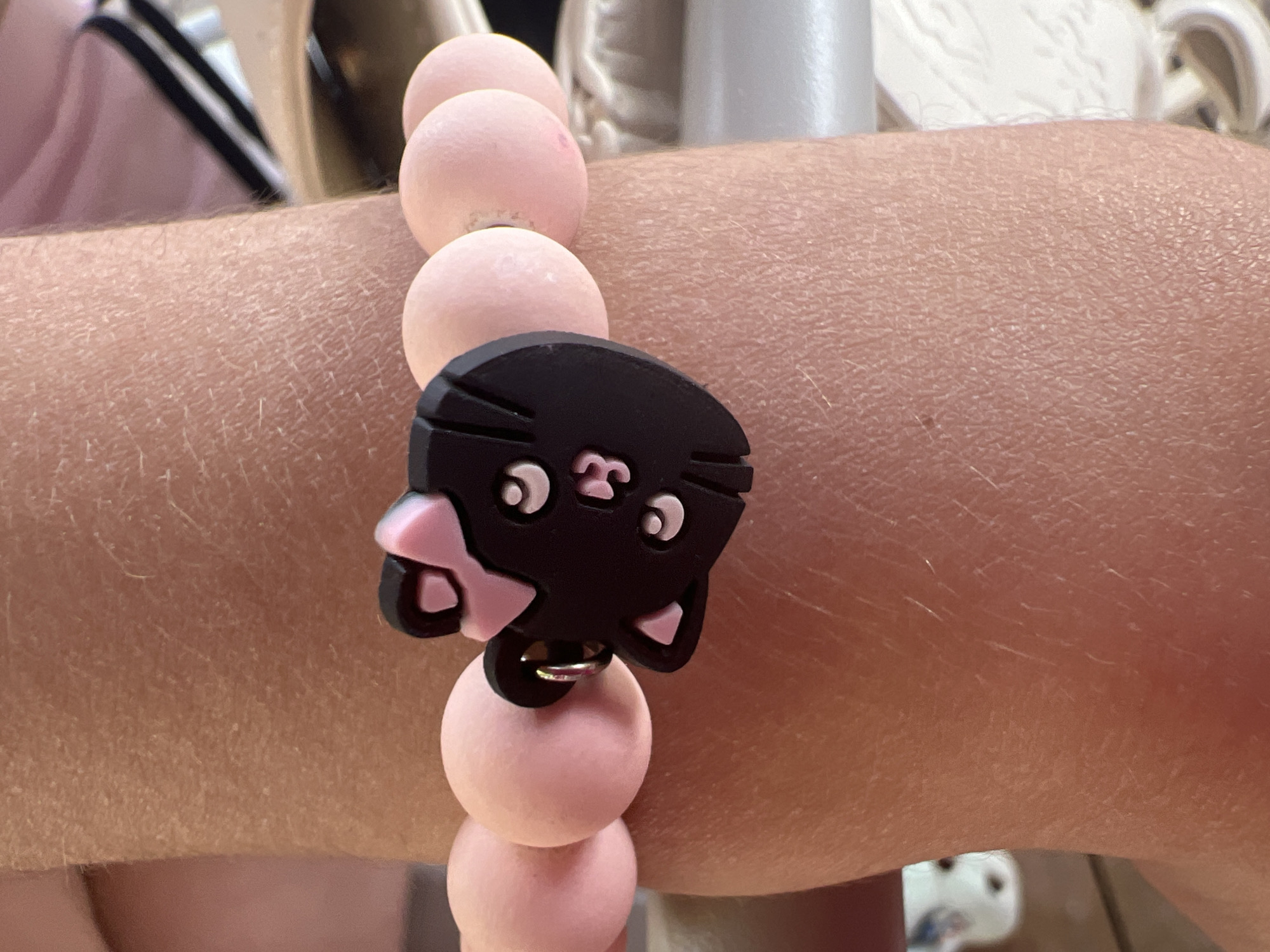 A close-up, eye-level shot features a black cat charm bracelet with a pink bow, worn by a person on their wrist. The bracelet is made of pink beads, and the cat charm has white eyes and a pink nose. The person's skin is visible, with fine lines and hairs. The background is blurred, but hints of light-colored surfaces and fabric can be seen. The image appears to be taken indoors.