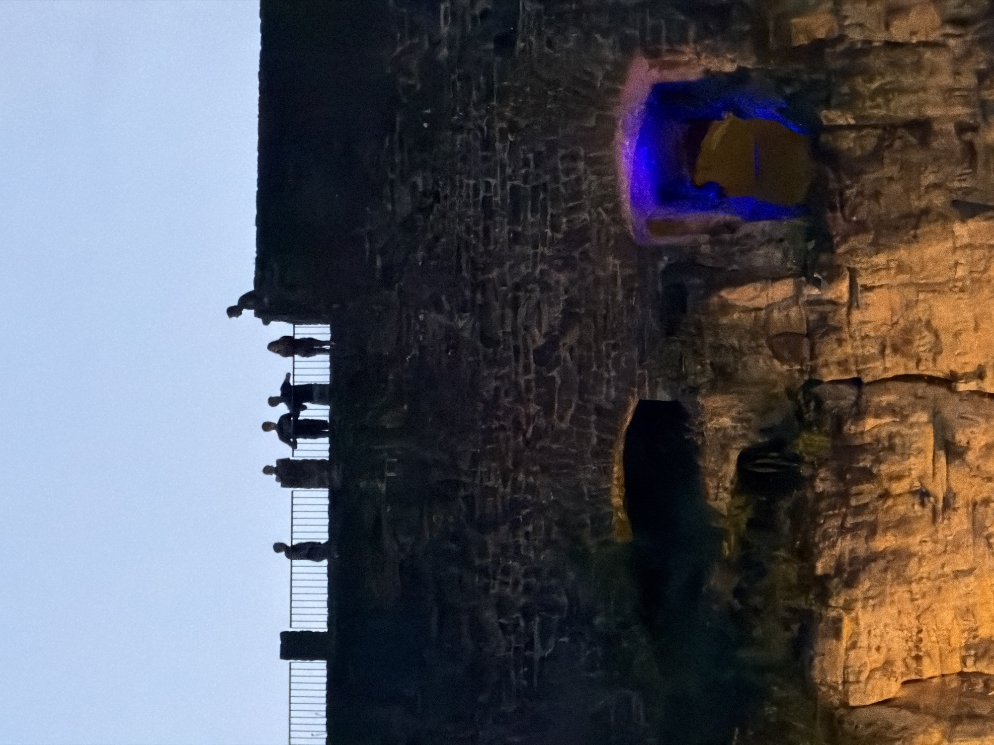 This image depicts a twilight scene with several silhouetted figures standing on a stone wall and railing overlooking a cliffside. The figures are grouped at various points along the walkway. Below the wall, the cliff face is visible, with a prominent archway illuminated by blue and yellow lights. The overall setting suggests an outdoor historical site or fortification. The location is the Bock Casemates in Luxembourg City, Luxembourg.