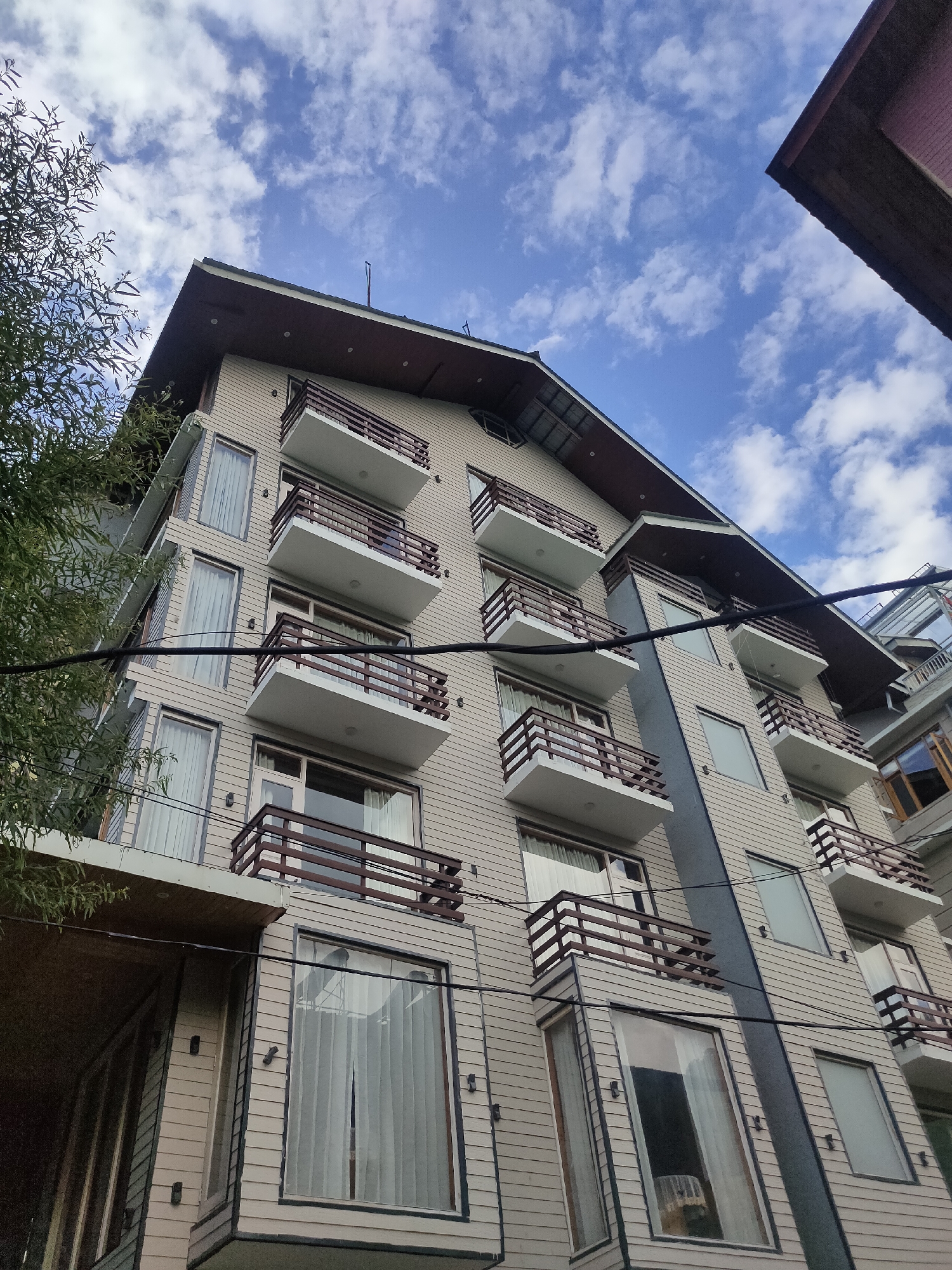 The image shows a modern, multi-story building with a light beige exterior, possibly made of wood or siding. The building features numerous balconies with dark brown railings, facing a cloudy blue sky. The balconies appear to be a prominent architectural element, each extending outwards from the building's facade. There are several windows visible, many of which are covered with light-colored curtains, suggesting they are residential or hotel rooms. A tree with sparse foliage is partially visible on the left side of the frame, adding a touch of nature to the urban landscape. Some electrical wires are strung across the building, indicating the presence of utilities. The overall impression is that of a well-maintained building in a city setting, likely in a mountainous region given the style and the cloudy sky. The location is Siumsa, India.