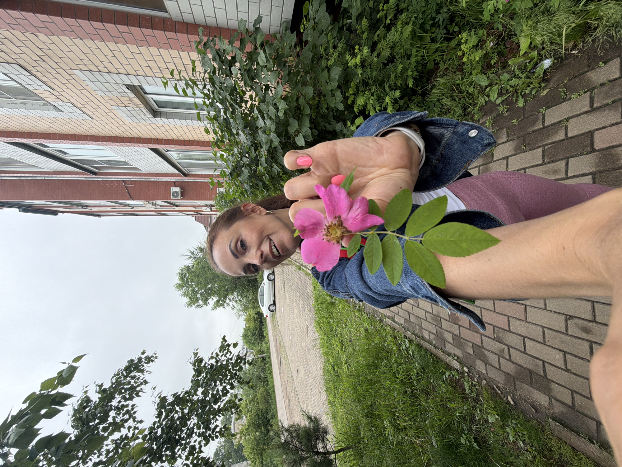 A woman with auburn hair, wearing a denim jacket and pink leggings, stands on a brick pathway. She is holding a pink flower with green leaves in her outstretched hand, close to the camera. Behind her, a multi-story building with a brick facade is visible. The environment suggests an urban setting in Birobidzhan, Russia, with trees and a paved parking area with a white car in the background. The sky is overcast.