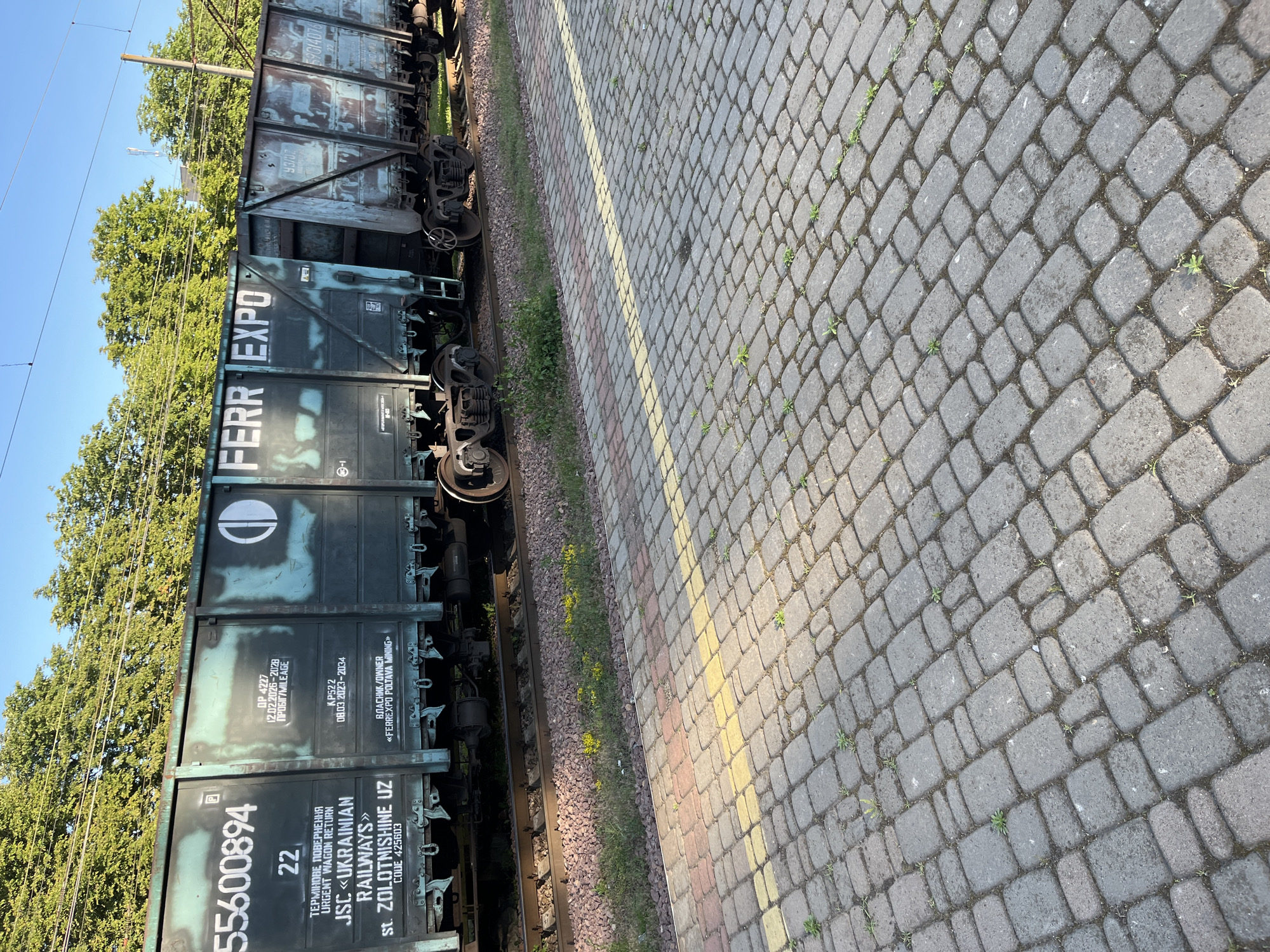 A freight train is parked on a railway track in an outdoor setting. The train cars are dark green and show signs of wear and tear. One car prominently displays the number "55600894" and "FERR EXPO" in white lettering. Other visible text includes operational details, dates, and company names like "JSC «UKRAINIAN RAILWAYS»". The train is situated next to a paved walkway made of grey and red cobblestones, with a yellow line demarcated on the pavement. In the background, a line of green trees forms a canopy against a clear blue sky. The scene appears to be a sunny day, with shadows cast across the paved walkway. There are no people visible in the image. The location is likely an industrial area or a train station in Kyiv, Ukraine, given the presence of Ukrainian railway information.