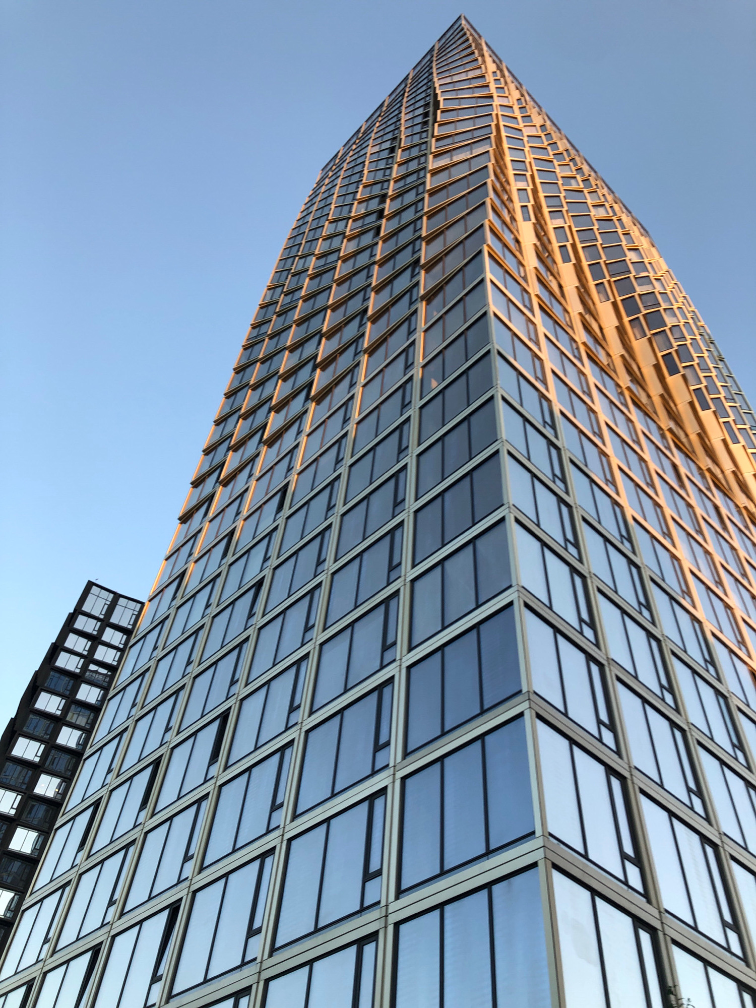 This is a low-angle, full-shot photograph of a modern skyscraper in Moscow, Russia, likely captured during sunset due to the warm, golden light reflecting off the building's facade. The skyscraper is characterized by its extensive glass curtain walls, with numerous windows arranged in a grid pattern. The building's design appears to have a slightly tapered or twisted form as it ascends, creating a dynamic visual effect.
To the left of the main skyscraper, another building is partially visible, also featuring a glass facade but with a darker, more angular construction. The sky is a clear, pale blue, transitioning to a warmer hue near the horizon, indicating the time of day.
There are no people visible in the image. The focus is solely on the architectural features of the buildings and the atmospheric lighting. The scene is clean and uncluttered, emphasizing the geometric lines and reflective surfaces of the contemporary urban architecture. The photograph highlights the scale and modernity of the structures within the Moscow cityscape.