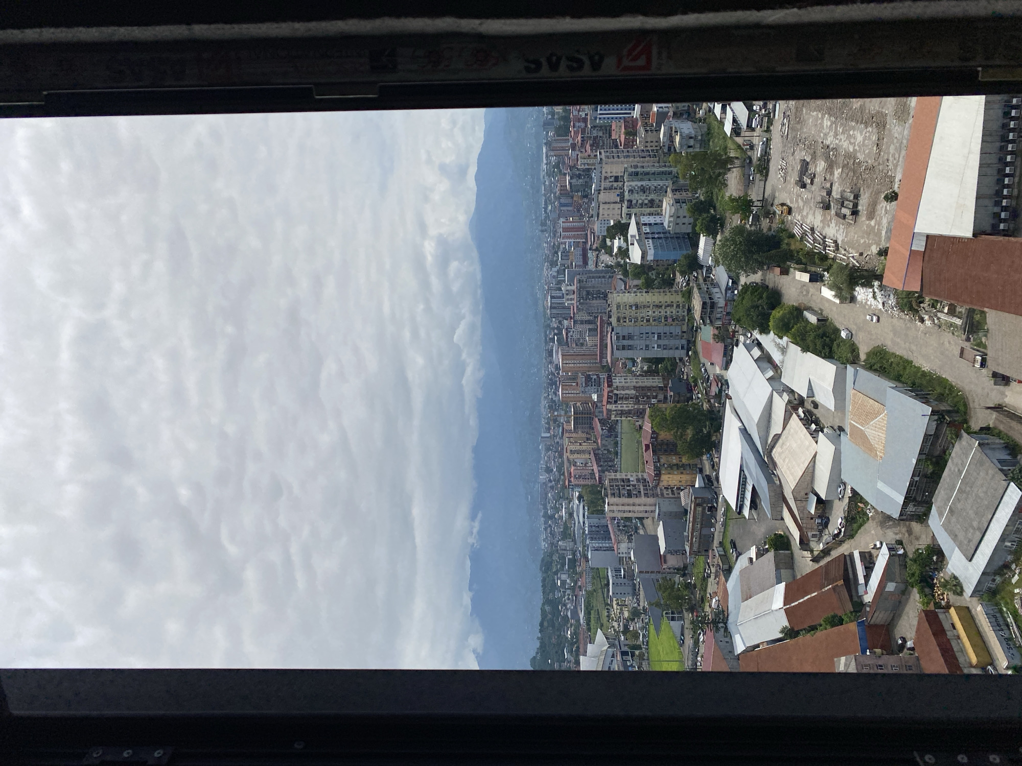 The image captures an aerial view of the city of Batumi, Georgia, looking towards a range of mountains under a cloudy sky. In the foreground, industrial buildings with corrugated metal roofs are visible, some appearing aged and weathered with rust. To the left, the dark frame of a window or balcony structure intrudes into the scene, with what appears to be graffiti or markings on its inner surface.
Beyond the industrial area, the cityscape unfolds with a mix of multi-story residential buildings, some with colorful facades, and lower-rise structures. A sports stadium or complex with a distinctive white roof is present on the left side of the mid-ground. Green spaces and roads with parked vehicles are interspersed throughout the urban landscape.
Further in the distance, the hazy silhouettes of mountains rise against the cloudy sky, forming a natural backdrop to the city. The overall impression is one of a developing urban environment with both industrial and residential zones, situated near a mountainous region. The lighting suggests it is daytime, likely overcast, which diffuses the sunlight. No people or active interactions are discernible in the image.