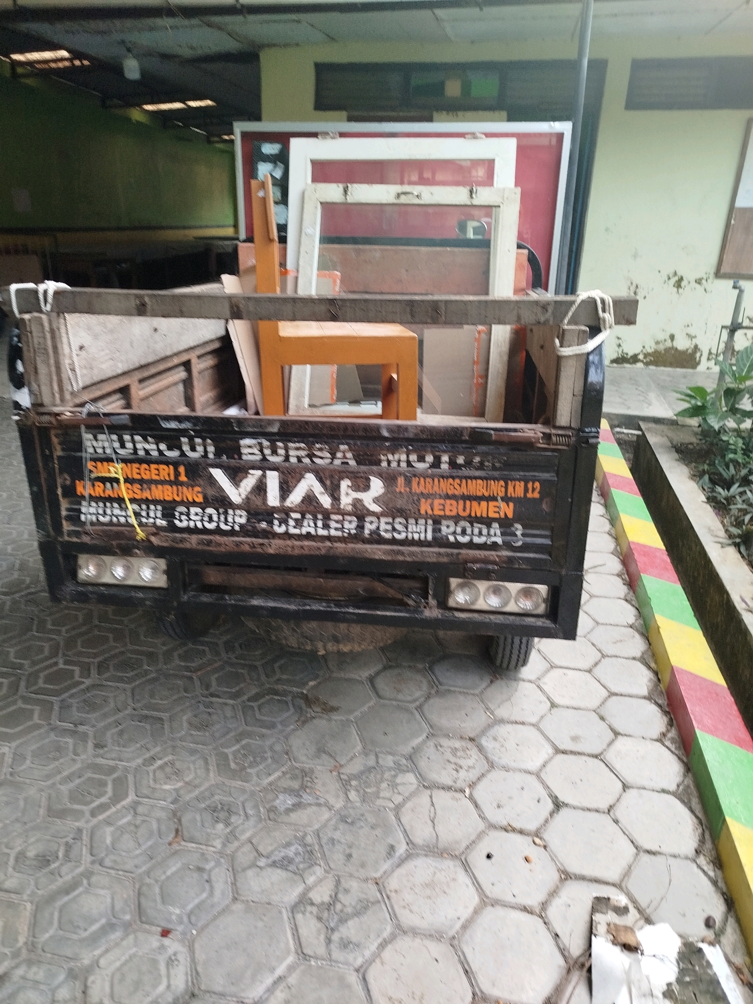 A black pickup truck with a wooden cargo bed is parked on a hexagonal paver surface outside. The truck bed contains wooden planks and a partial view of an orange chair. Text is visible on the side of the truck bed, including "MUNCUI", "BURSA MOTOR", "SMK NEGERI 1", "KARANGSAMBUNG", "VIAR", "JL. KARANGSAMBUNG KM 12", "KEBUMEN", "MUNCUL GROUP", and "DEALER PESMI RODA 3". The truck has three brake lights on the rear. A wall with faded paint and some green foliage is visible in the background. The scene appears to be in Kedungwaru, Indonesia, given the presence of place names like Karangsambung and Kebumen on the truck.