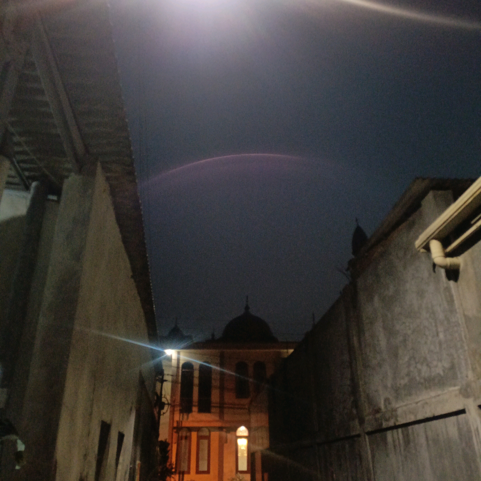 A low-angle, nighttime photograph taken in Salatiga, Indonesia, captures a street scene with an illuminated building in the background and surrounding structures. The sky above is a deep, dark blue, with a faint, purplish arc across the upper portion, possibly a distant light phenomenon or artifact of the camera lens. In the center of the frame, a building with architectural elements suggesting a mosque or similar place of worship is visible, featuring domed roofs and arched windows. The building is lit from within, casting a warm orange glow. The foreground is dominated by the textured walls of buildings on either side, creating a narrow alleyway effect. The left side of the image shows a wall with a corrugated roof structure extending over it. The right side features a concrete wall with a drainage pipe. Light streaks, likely from artificial lighting, cut across the scene, adding to the nocturnal atmosphere. No people are visible in the photograph.