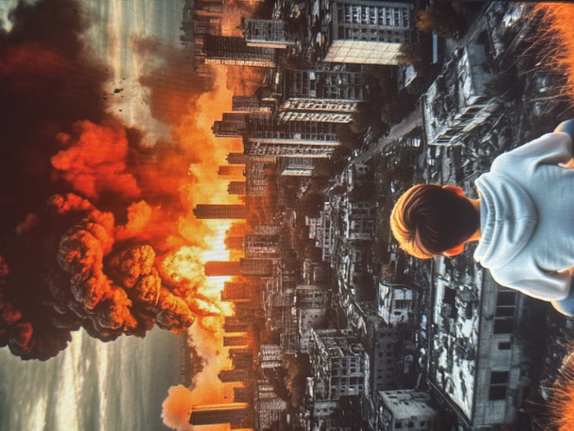 A wide shot depicts a city engulfed in a massive explosion, viewed from behind a person sitting on a hill overlooking the scene. The explosion is a towering cloud of fire and smoke, with intense oranges and reds dominating the sky. Below the explosion, a cityscape of buildings, some appearing damaged or destroyed, stretches out. The explosion's light casts a warm glow over the urban environment. The person in the foreground is facing away from the camera, their back to the viewer, appearing to observe the catastrophic event. The overall atmosphere is one of destruction and perhaps a sense of awe or despair.