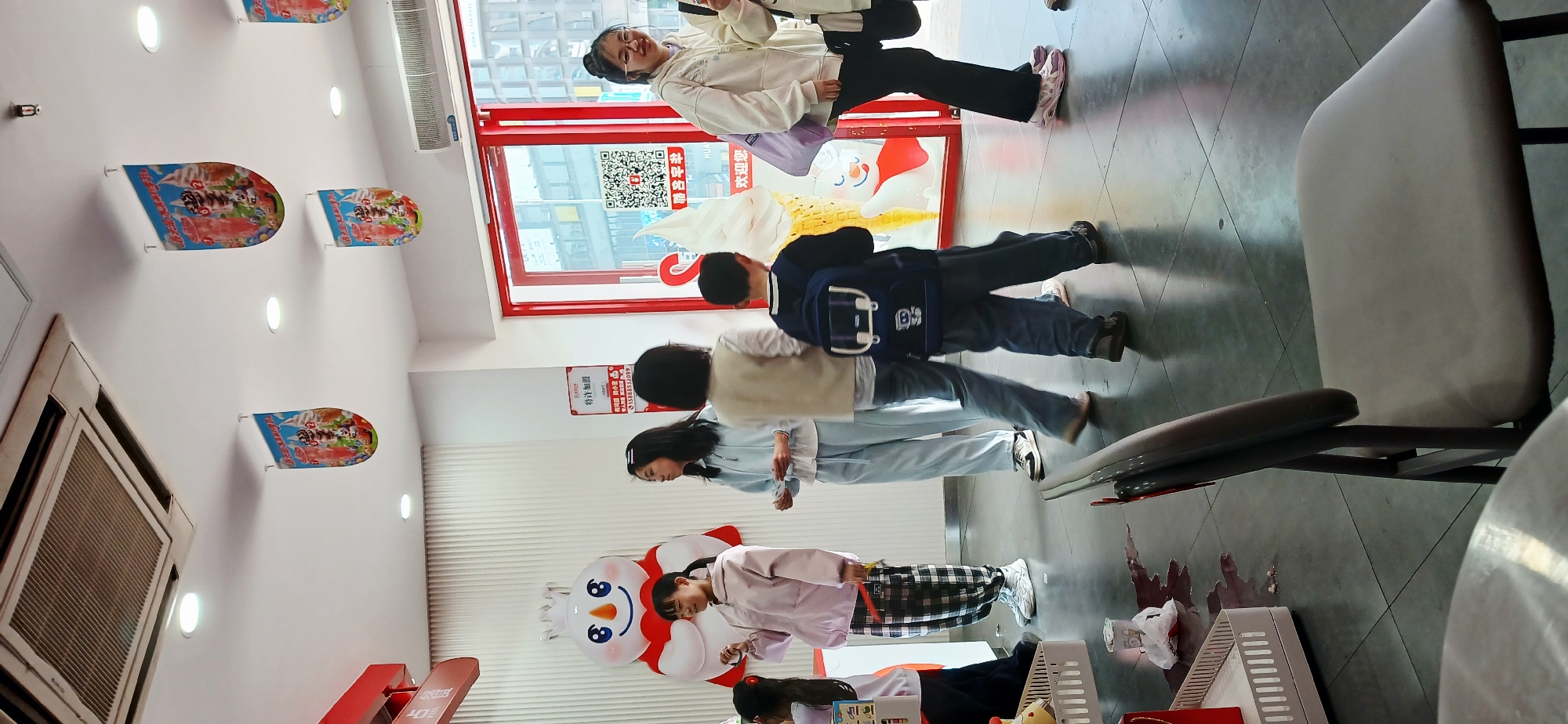 The photo shows the interior of a Mixue store in Jiulongpo District, China. Several people, mostly children, are present. From left to right: a girl is reading from a desk. To her right is a snowman-shaped standee. A girl in a checkered skirt is standing nearby. A young woman with dark hair and a girl in a light blue outfit are standing beside each other. A boy with a backpack is facing away from the camera, followed by a young woman with white jacket facing towards the street window. The floor is tiled. A chair is visible on the right. There are advertisements hanging on the wall and an air conditioning unit. Outside the window, buildings are visible. A red-framed glass door has a large ice-cream shaped logo on it.