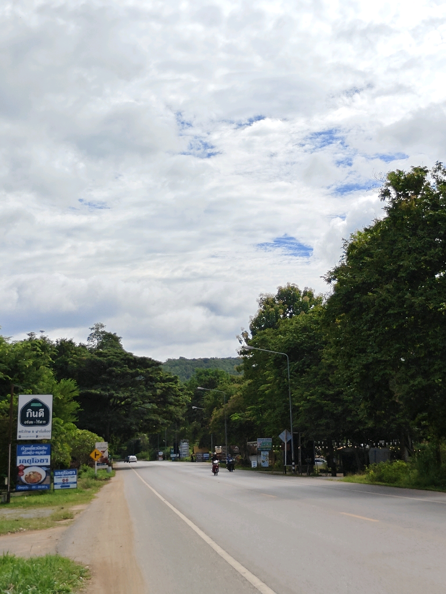 This is a vertical, eye-level, medium shot of a road in Khao Yai, Thailand. The road is paved and runs from the bottom right of the frame towards the upper left. There are cars and motorcycles on the road. On the left side of the road, there is a dirt shoulder with grass and signs. The signs are in Thai and appear to be advertisements or directional information. Behind the signs, there are trees and foliage. On the right side of the road, there are more trees and foliage, with some buildings visible in the distance. The sky is cloudy with patches of blue. The overall impression is of a rural or suburban area in Thailand.