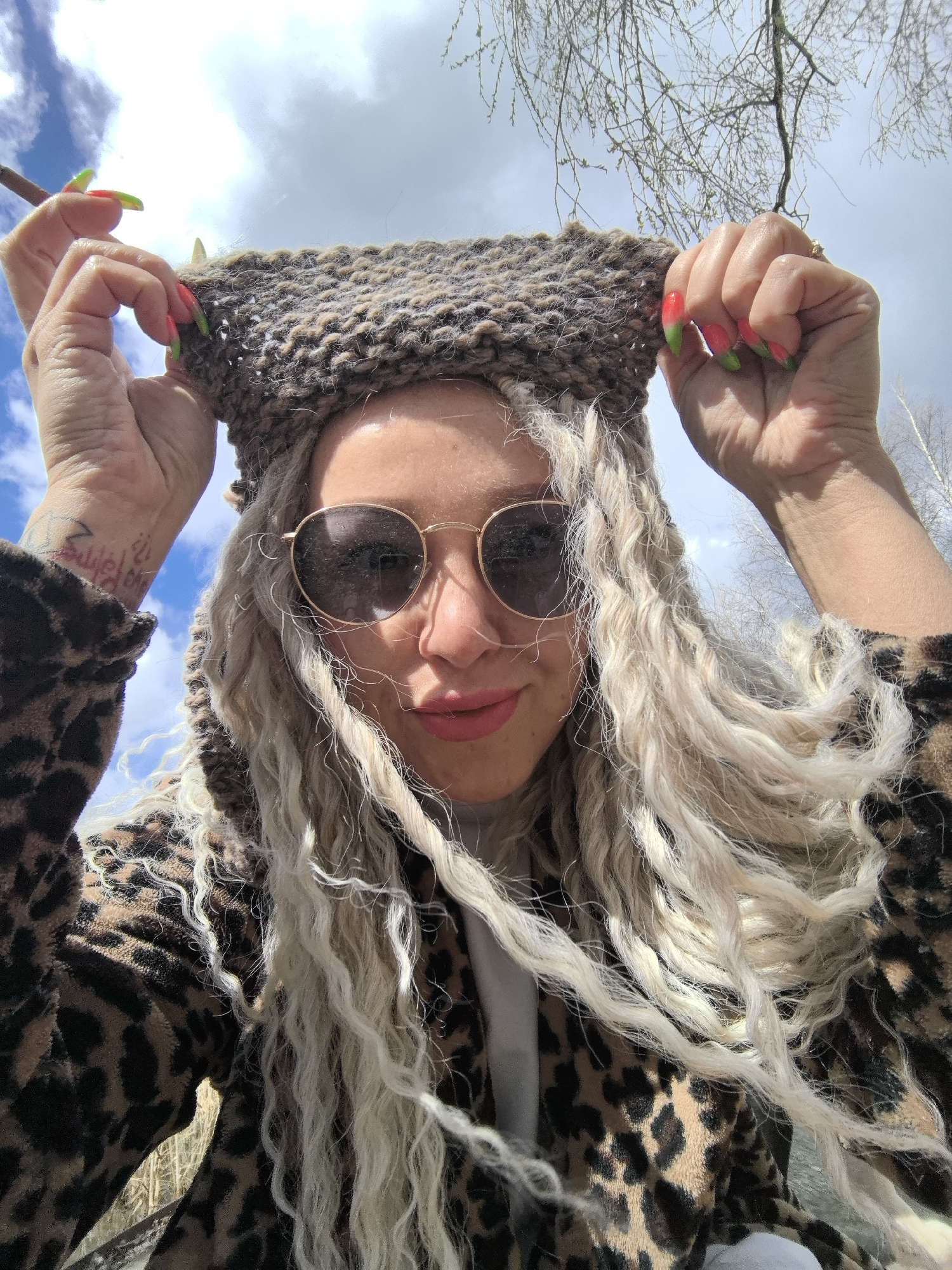 A woman with long, curly blonde hair, possibly dreadlocks, is captured in a close-up, upward-angled shot against a bright, cloudy sky in Noviy, Russia. She wears round, gold-rimmed sunglasses with dark lenses and a brown and black leopard print garment. Her lips are slightly parted in a gentle smile, showing pink lip color. She holds up a piece of brown, chunky knitted fabric, with knitting needles visible in her hands. Her fingernails are long and painted with a distinctive red/pink to green gradient design. On her left wrist, a tattoo with indistinct script or symbols is visible. The background features a bright blue sky with white clouds, and bare tree branches without leaves are visible on the right, suggesting late winter or early spring. The overall lighting indicates daytime. The woman appears to be playfully interacting with her knitting project.