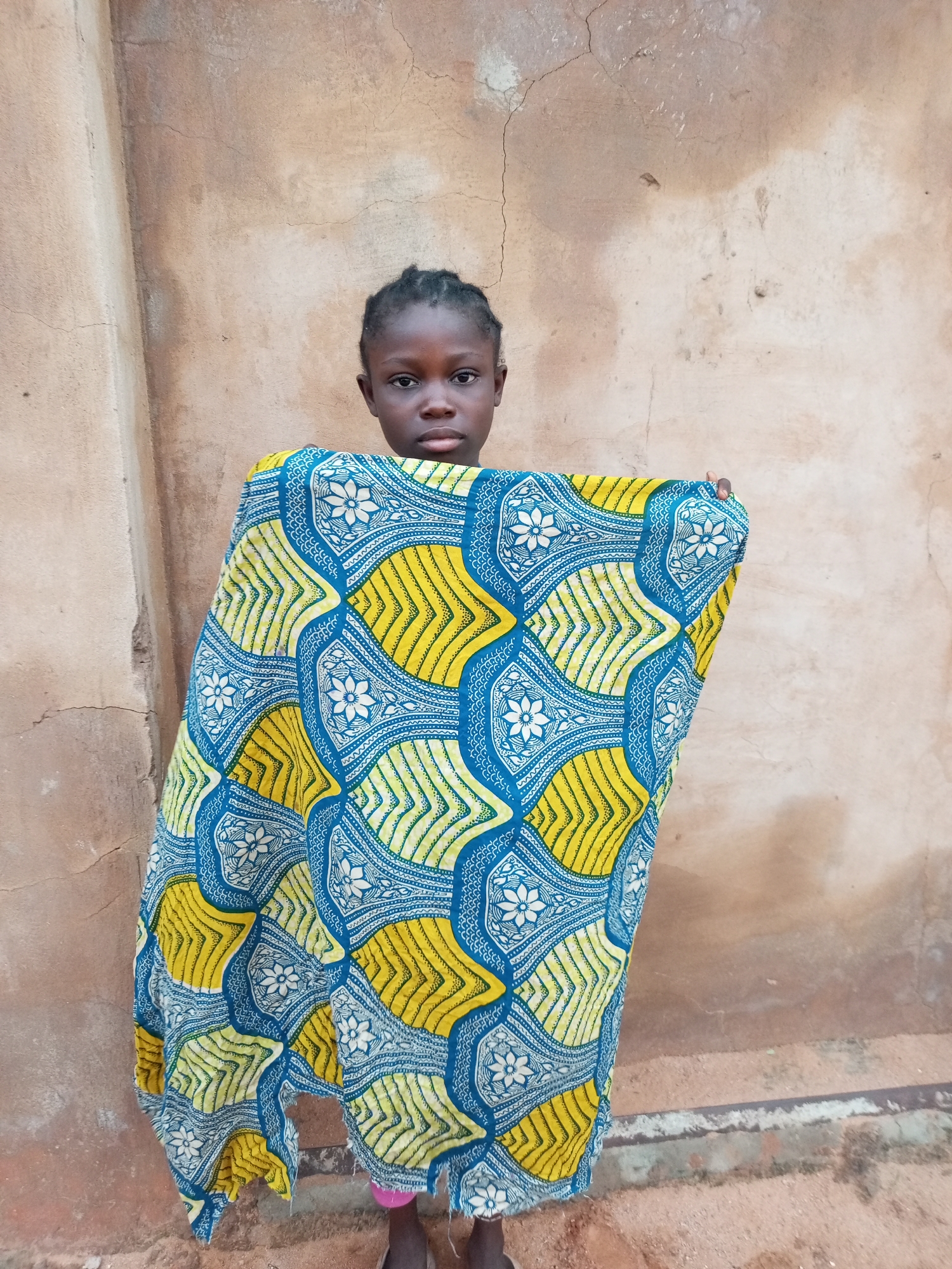 A young girl with braided hair stands in front of a textured, tan-colored wall. She is wrapped in a vibrantly patterned fabric that features yellow and blue designs, including floral motifs and geometric shapes. The fabric is draped over her shoulders and hangs down to her waist, obscuring most of her body. The wall behind her has visible cracks and imperfections, suggesting an outdoor setting. The ground in front of the wall appears to be dirt or sand.