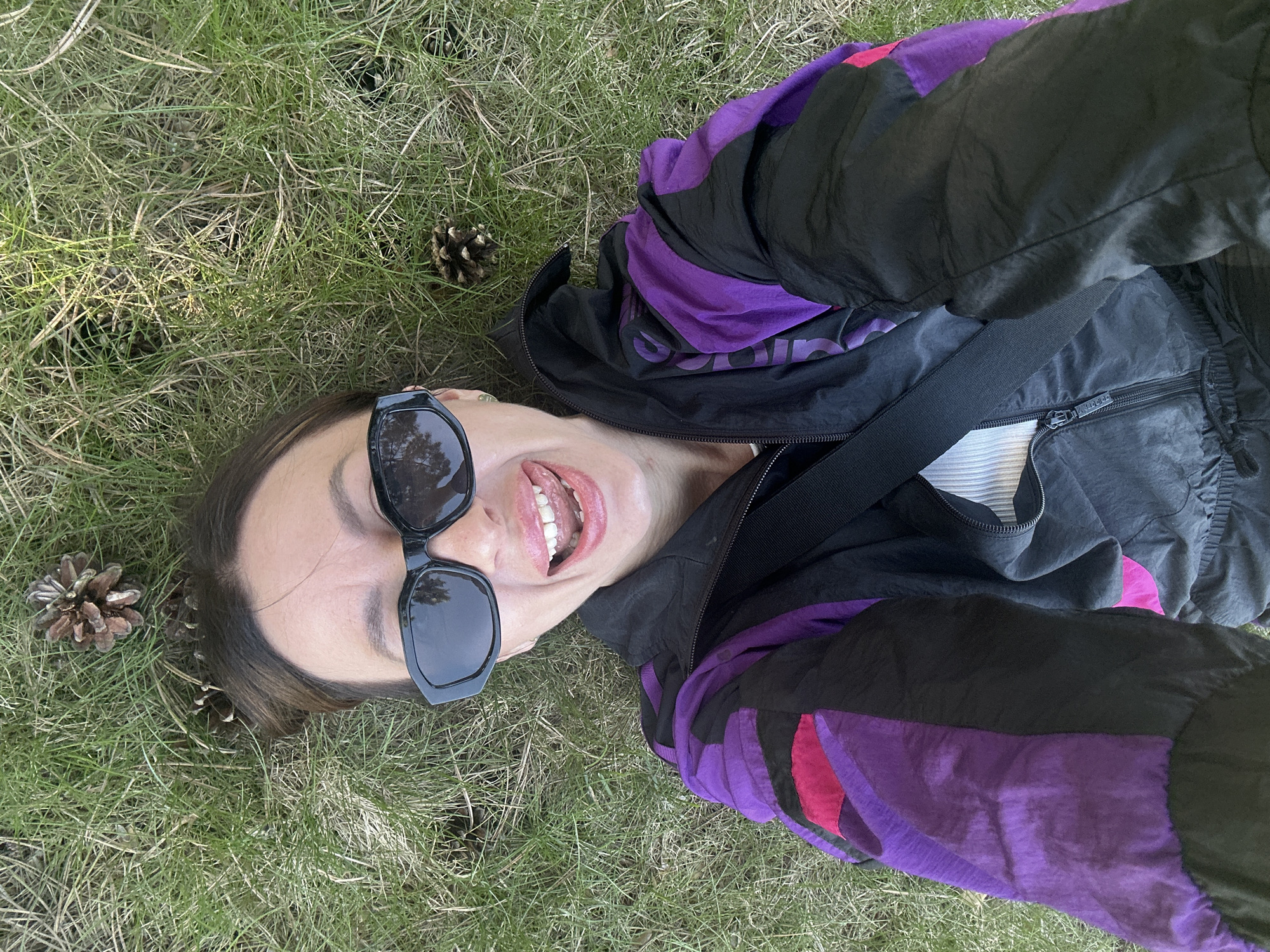 A woman is lying on grass, looking at the camera with her mouth open and wearing sunglasses. She is wearing a jacket that is black and purple. There are pine cones on the grass near her head. The location is not discernible from this image.